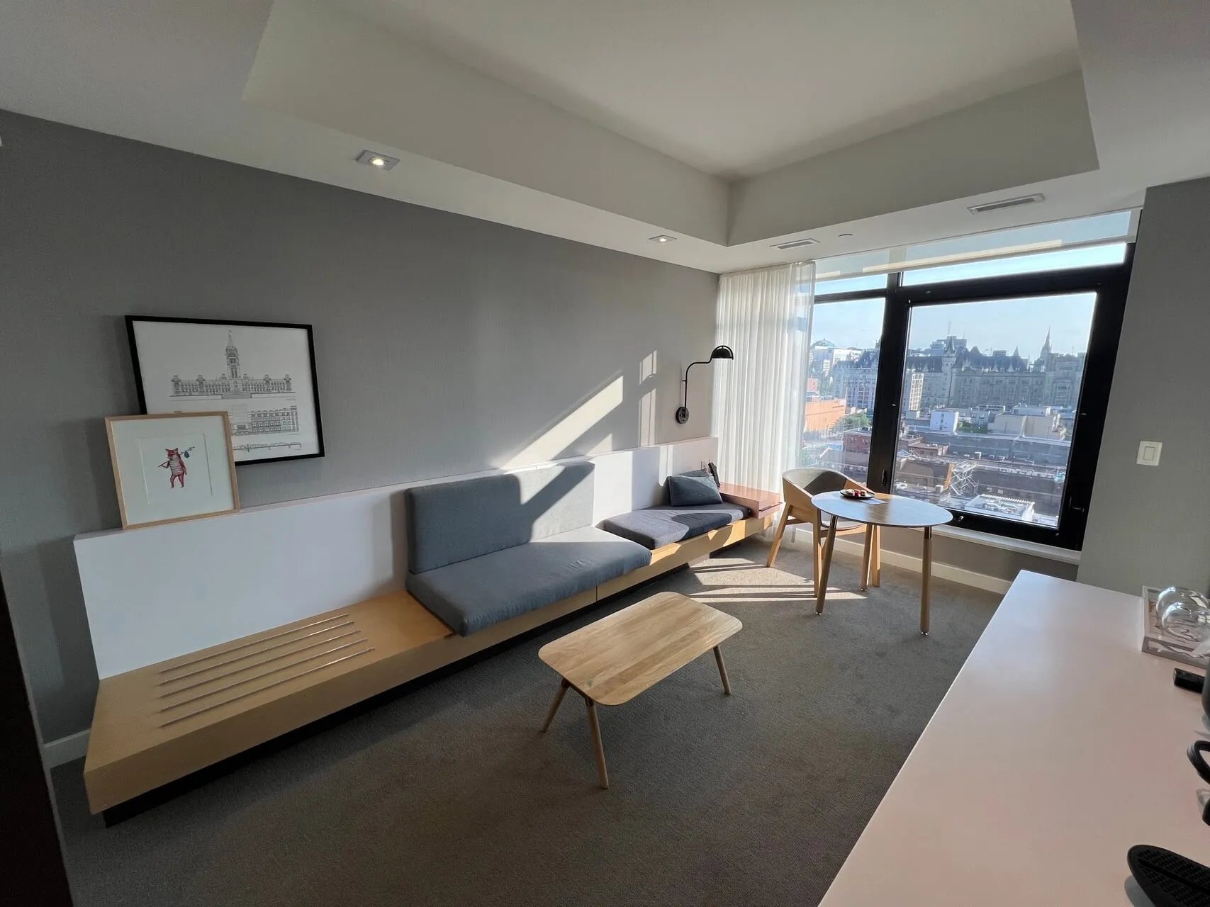 Modern hotel suite living room with gray sofa, wooden furniture, and city view of Ottawa skyline from floor-to-ceiling windows
