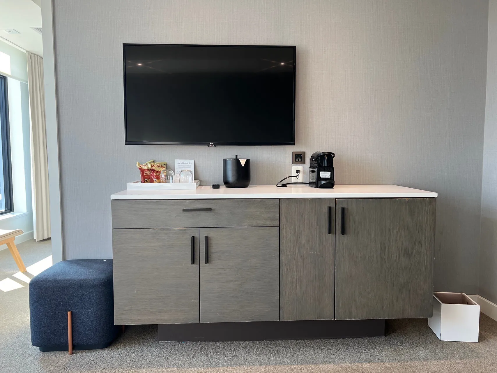 Entertainment center with wall-mounted TV, gray cabinetry, coffee maker, and blue ottoman in a modern hotel room