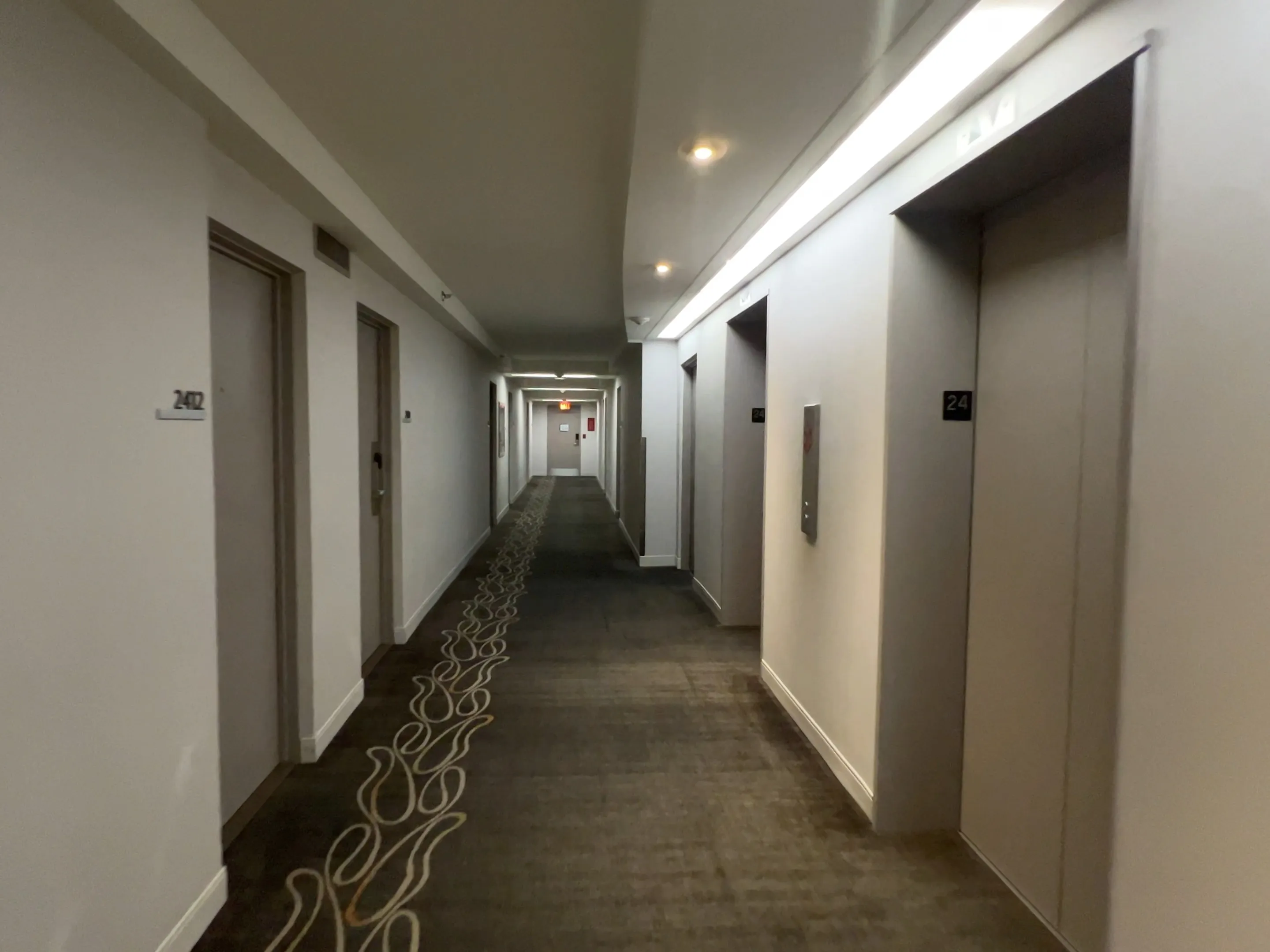 Hotel corridor with cream-colored doors, dark carpet, and modern LED ceiling lighting leading toward elevators.
