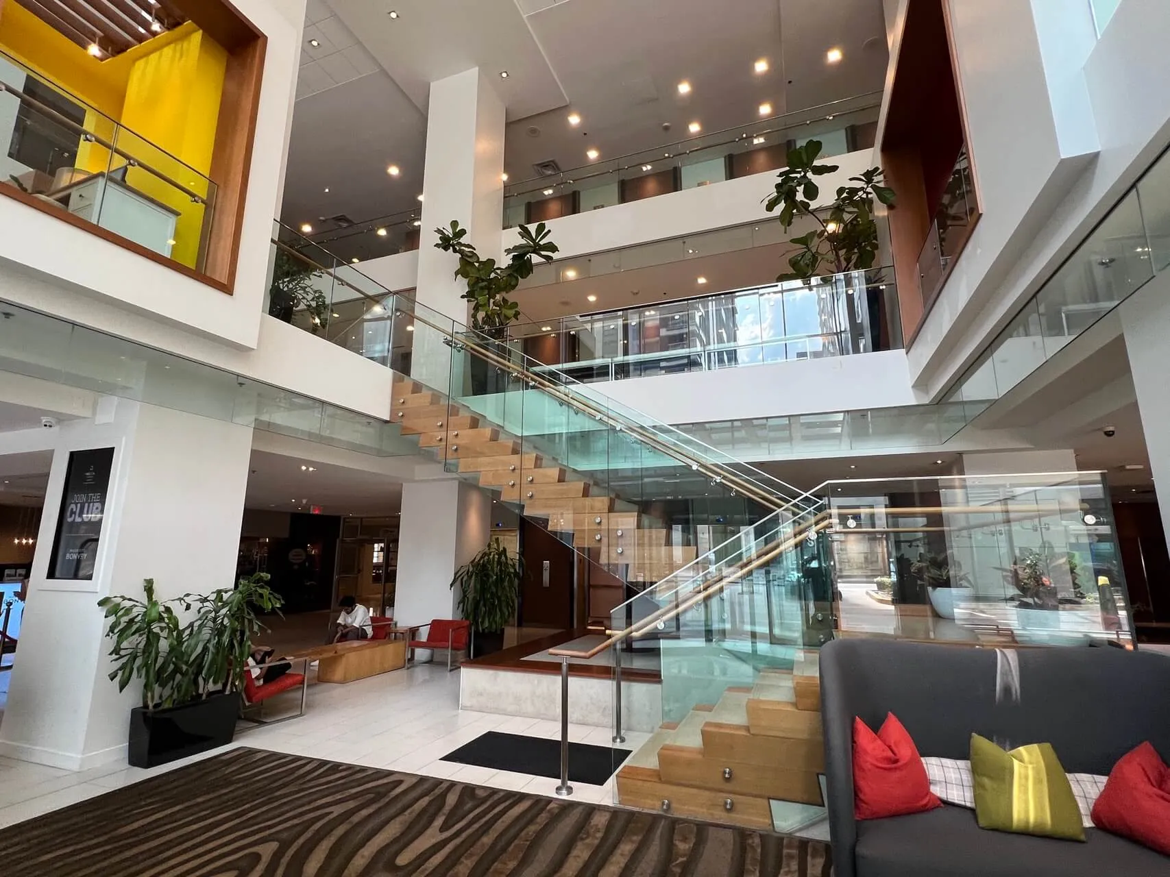 Multi-level modern hotel lobby with wooden staircase, glass railings, and contemporary furnishings.