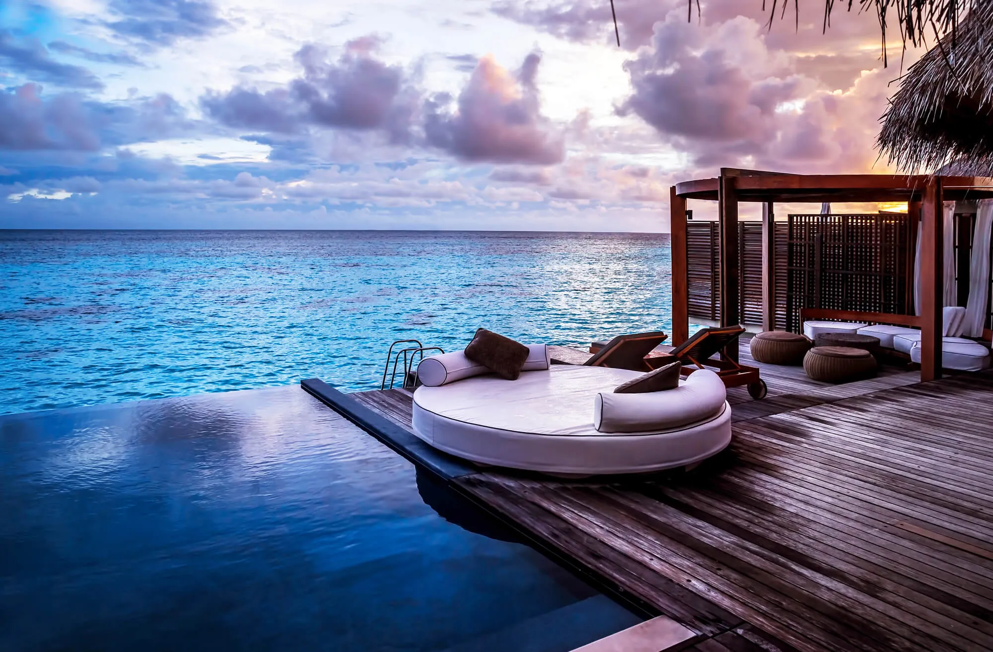 Overwater villa deck at sunset with circular daybed lounge, wooden boardwalk, and tropical ocean view
