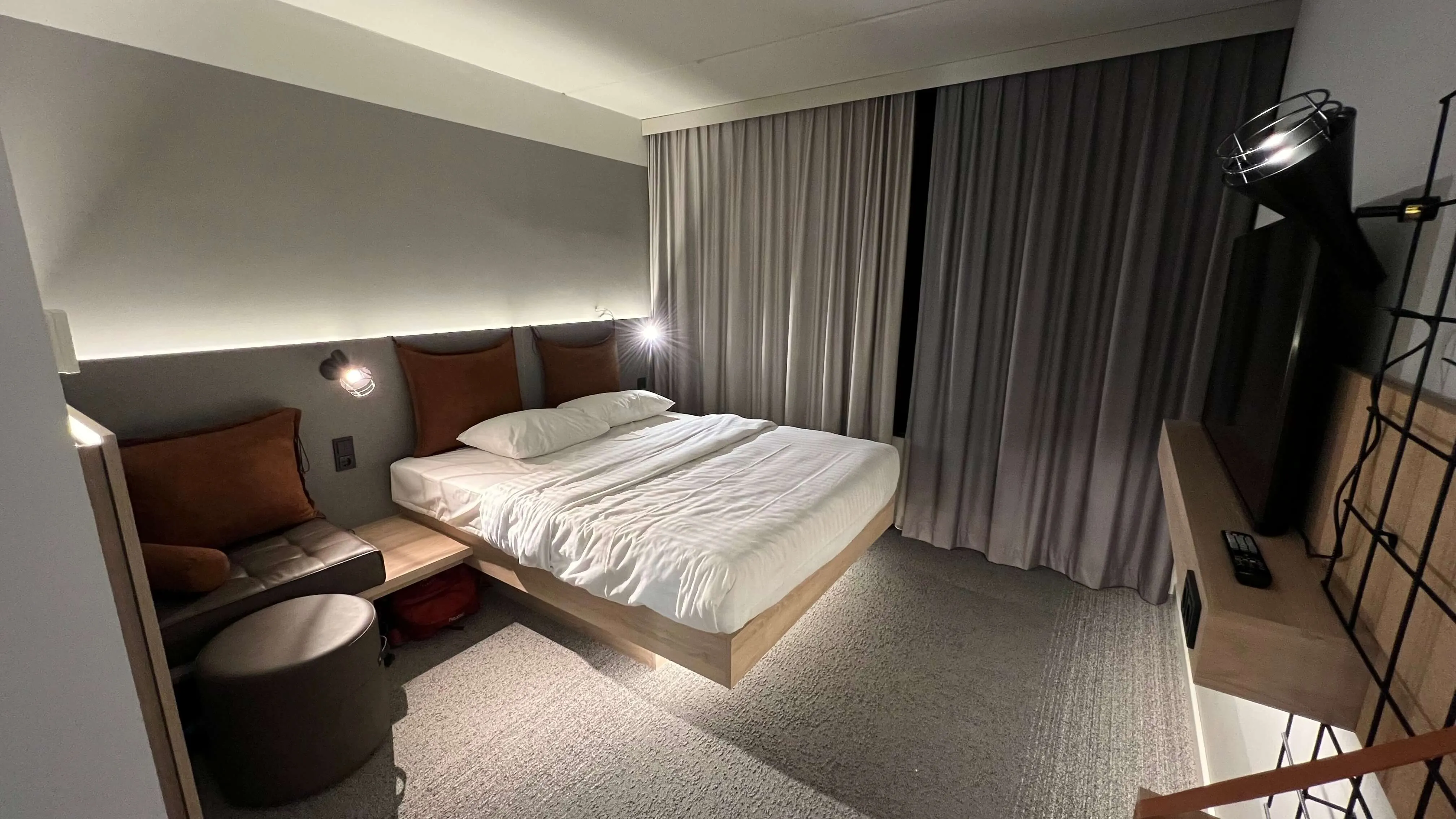 Modern queen bedroom with ambient headboard lighting, rust-red accent pillows, and floor-to-ceiling curtains at Moxy Frankfurt City Center