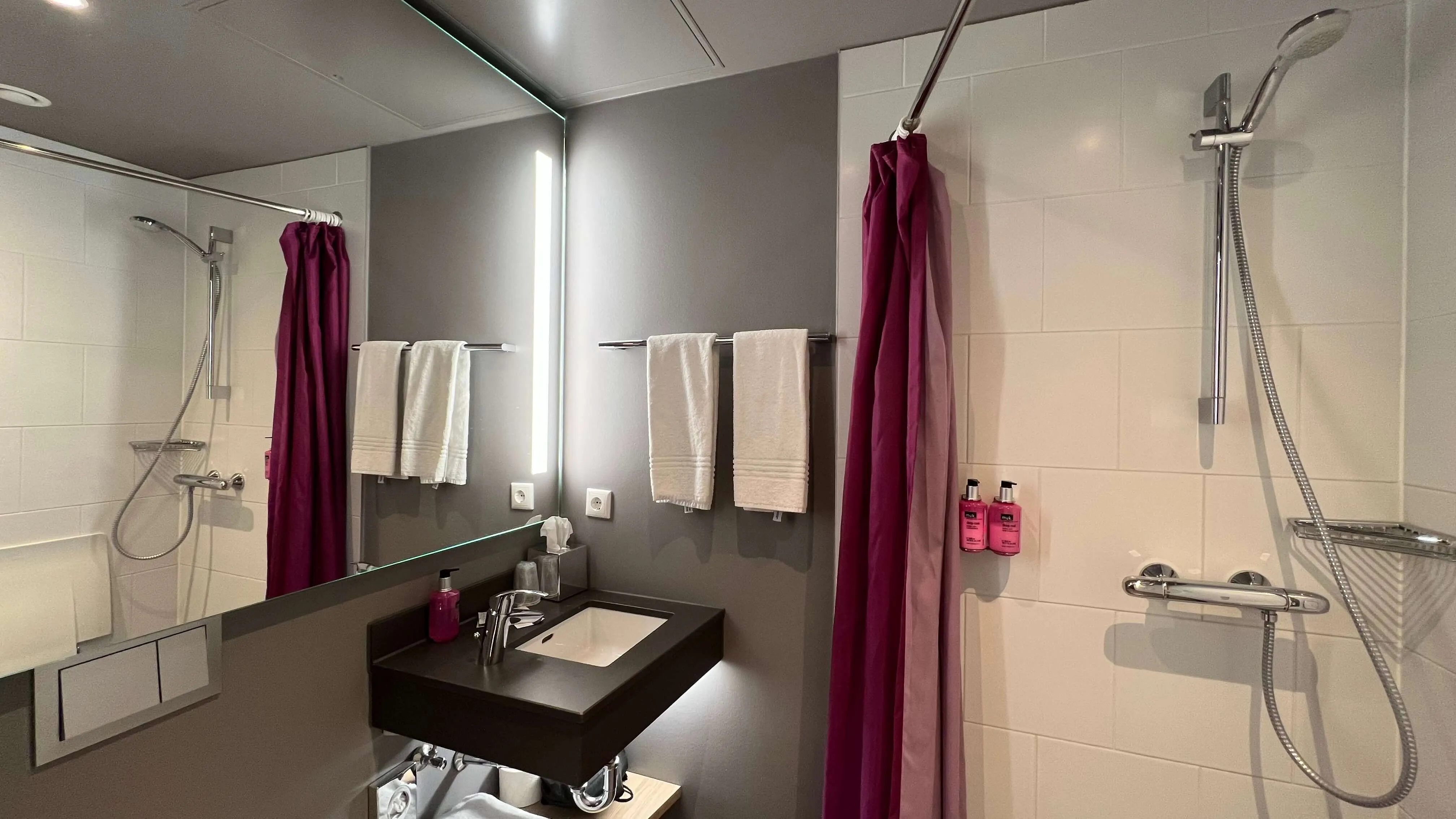 Modern hotel bathroom with black floating sink, illuminated mirror, burgundy shower curtains, and walk-in showers at Moxy Frankfurt City Center