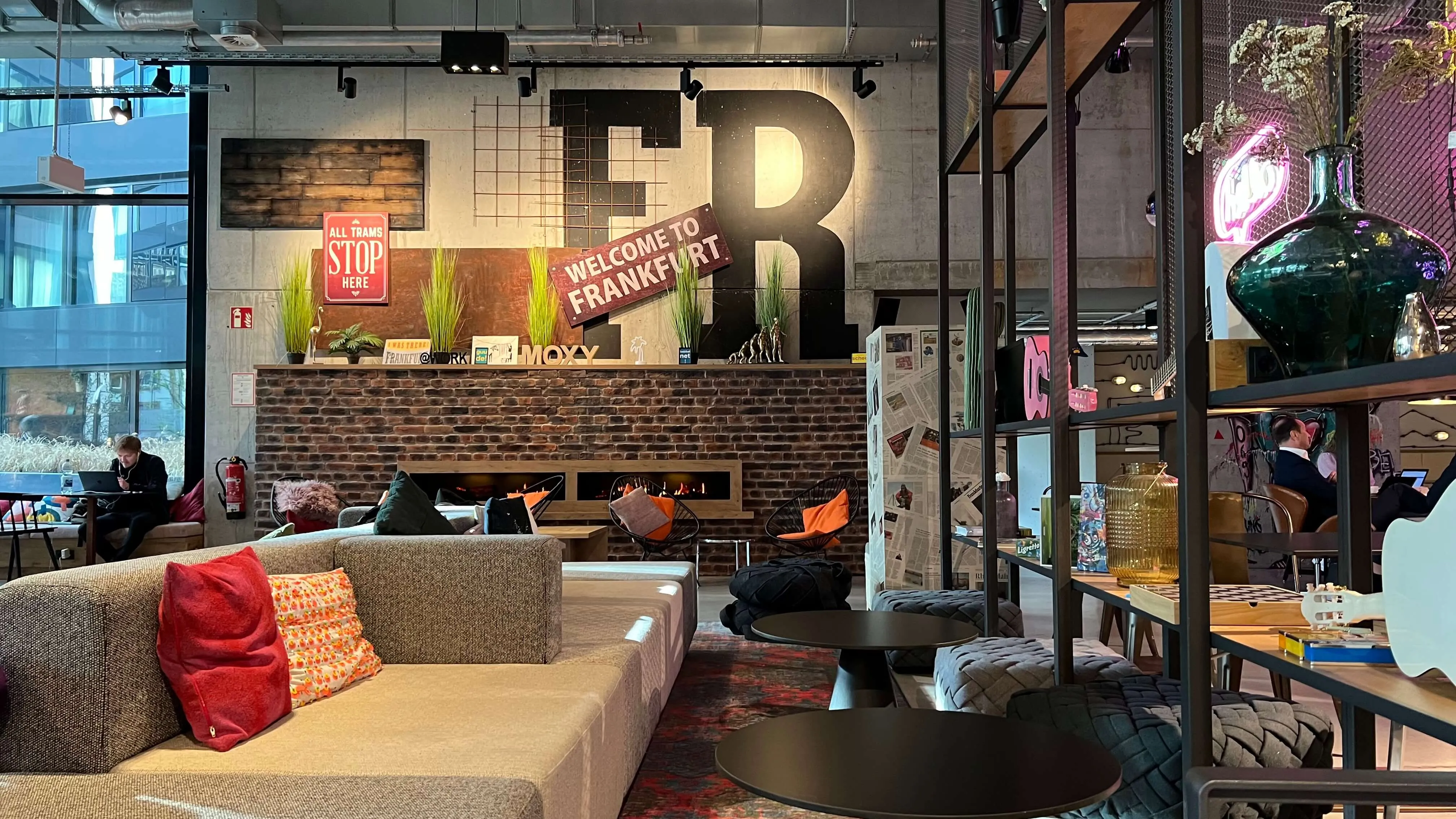 Moxy Frankfurt City Center lobby with tan seating, red accent pillows, and large illuminated 'FR' signage above brick counter