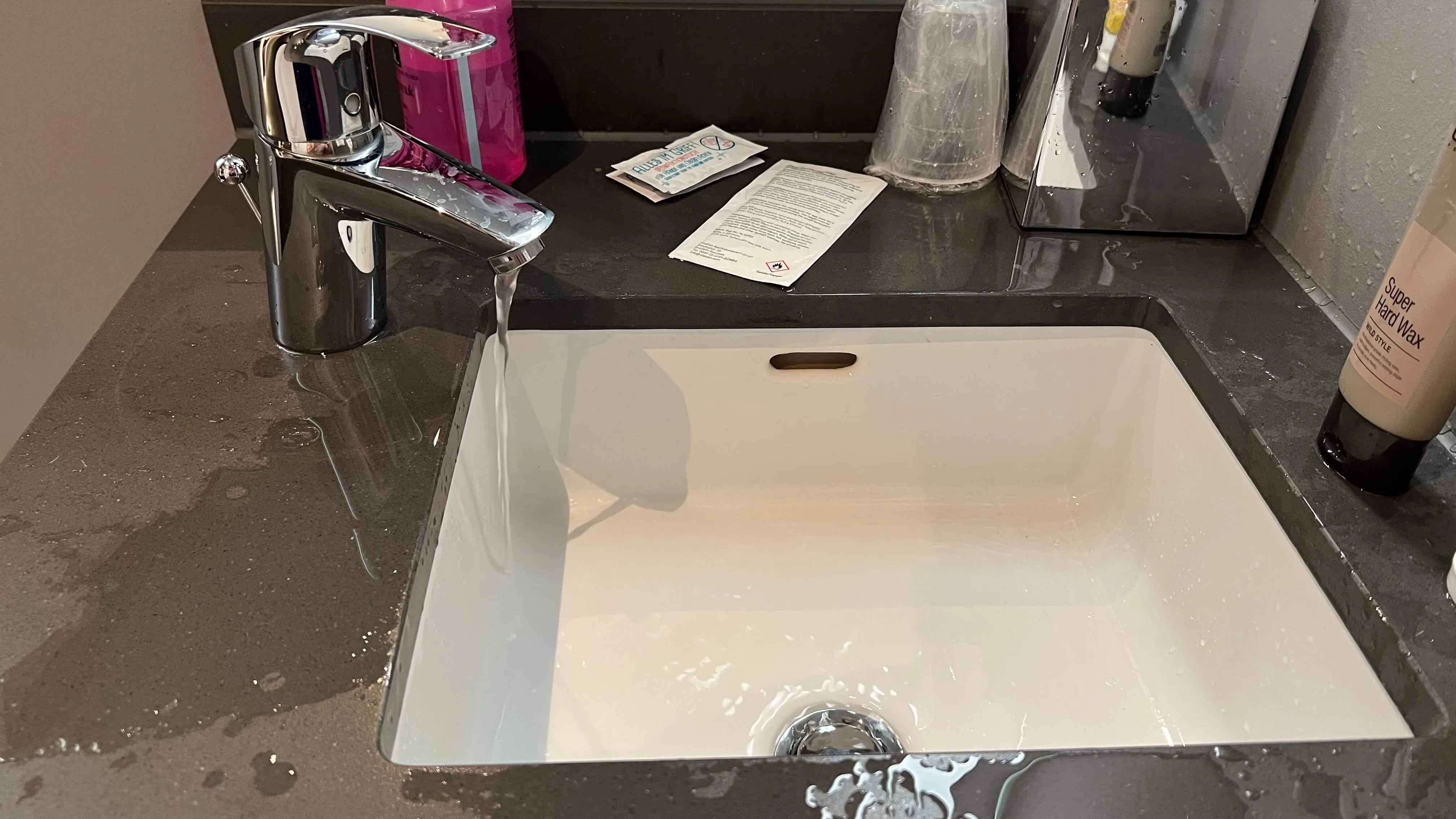 Modern washstand with rectangular white sink, chrome faucet, and dark countertop with toiletries and amenities at Moxy Frankfurt City Center