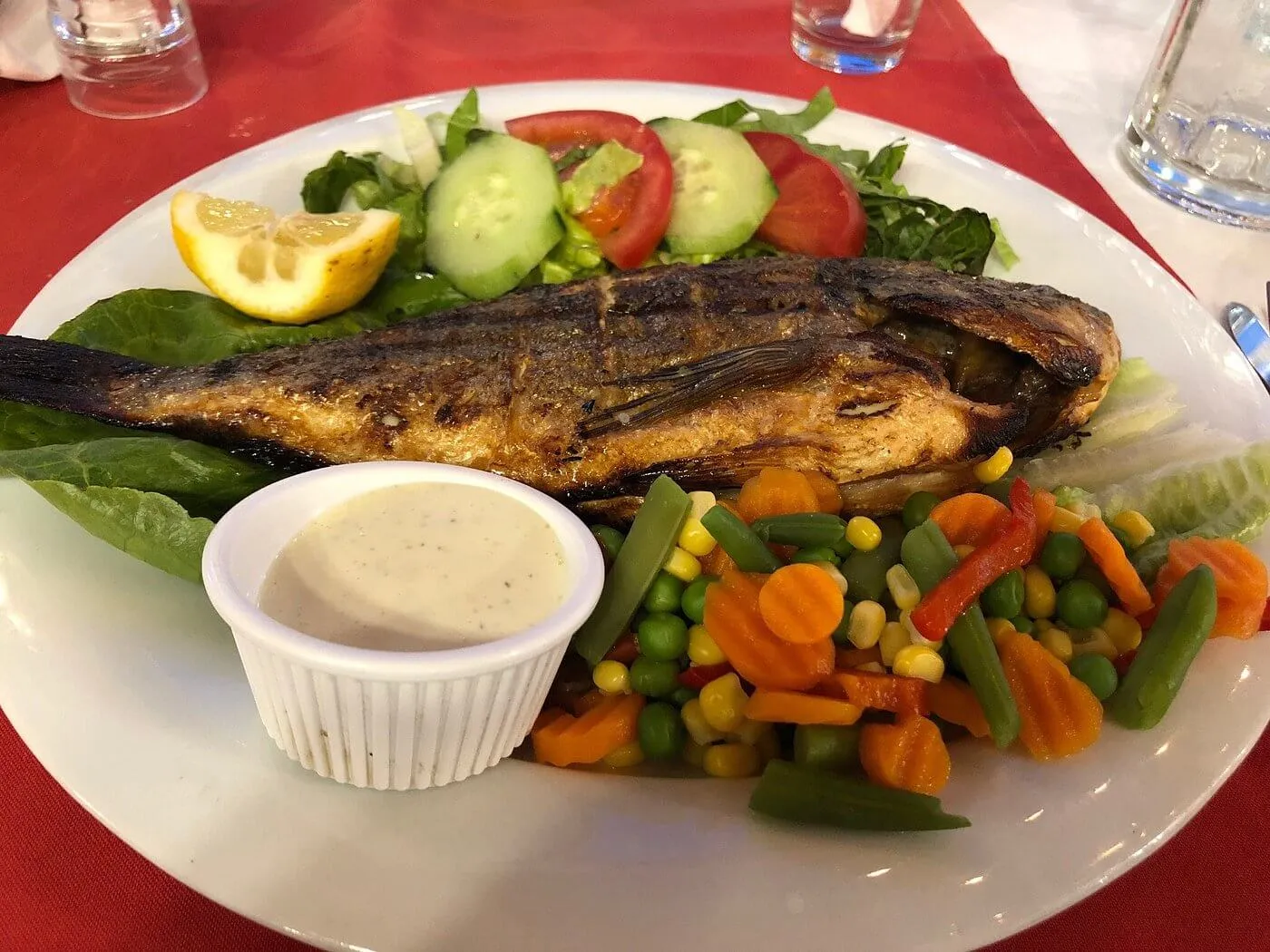 Grilled whole fish with lemon, cucumber, tomato salad, mixed vegetables, and white sauce on white plate at red tablecloth restaurant