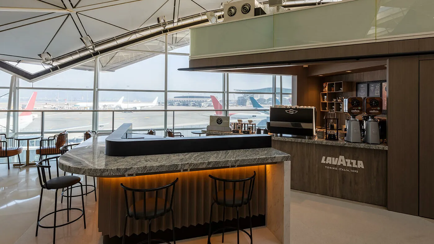 Plaza Premium lounge coffee bar with Lavazza branding, airfield view through floor-to-ceiling windows, black bar stools, and aircraft parked at gates