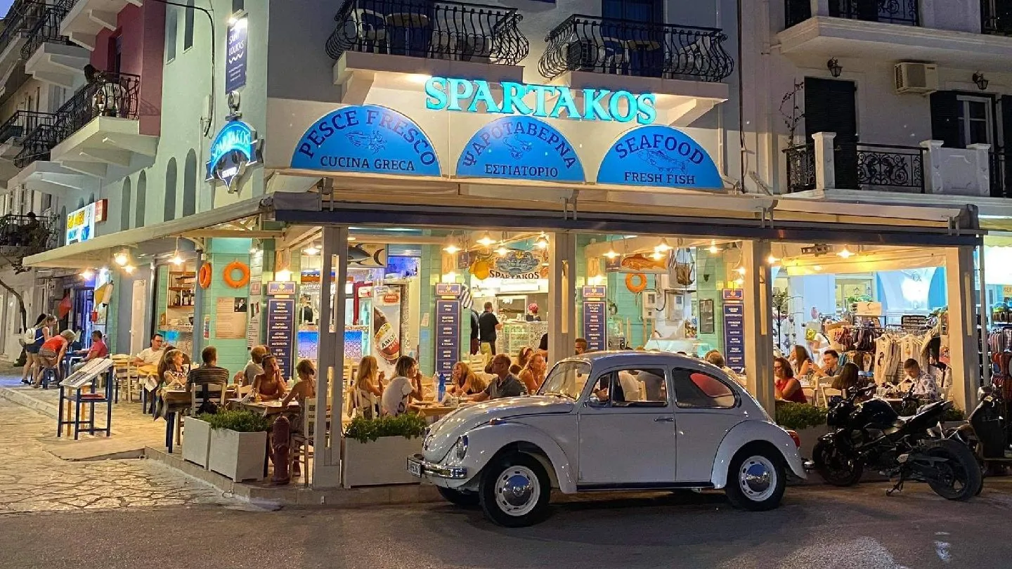 Spardakos seafood taverna storefront at night on a Greek street with blue signage and a Volkswagen Beetle parked in front