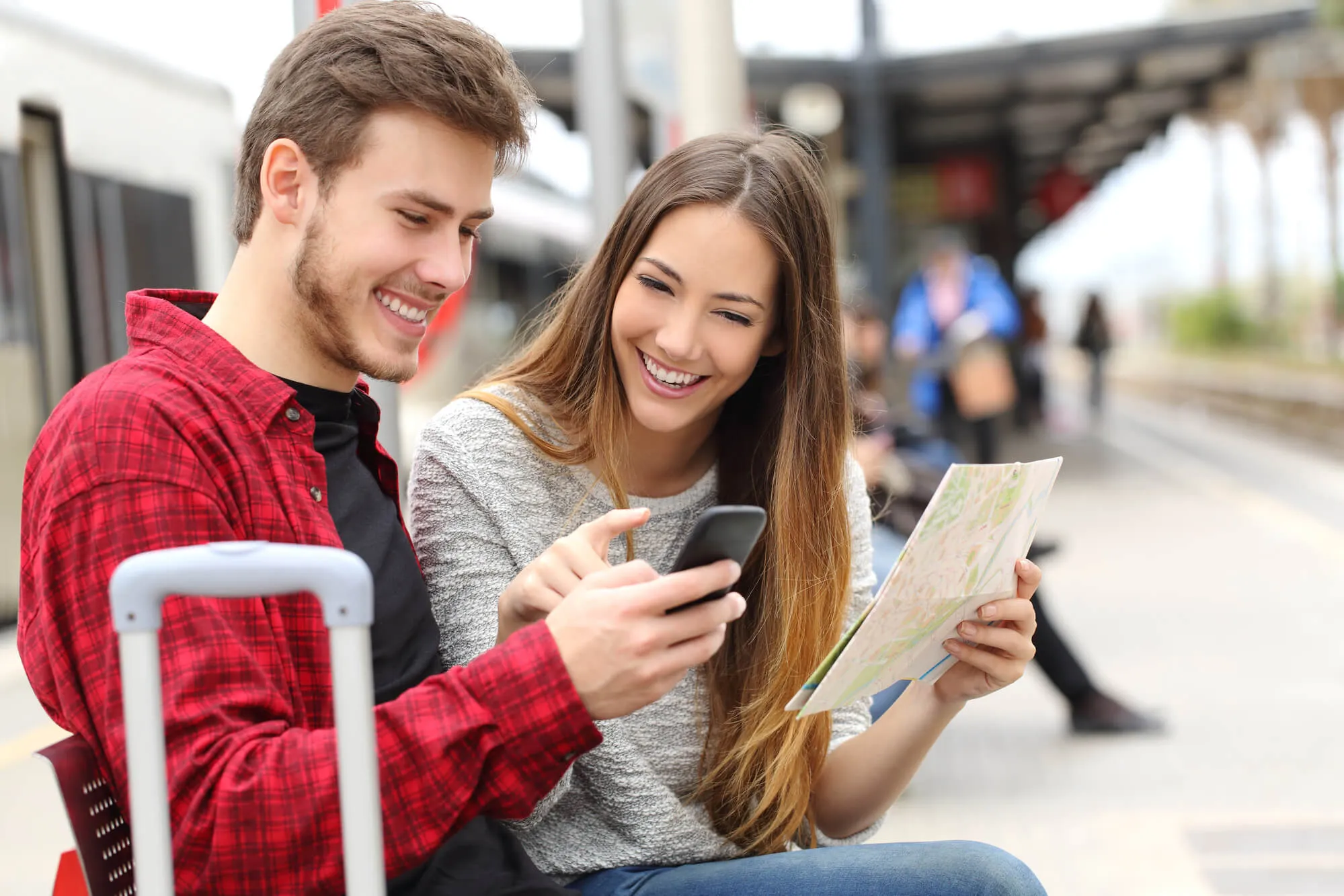 Young couple consulting a map and smartphone at a transit station with luggage.