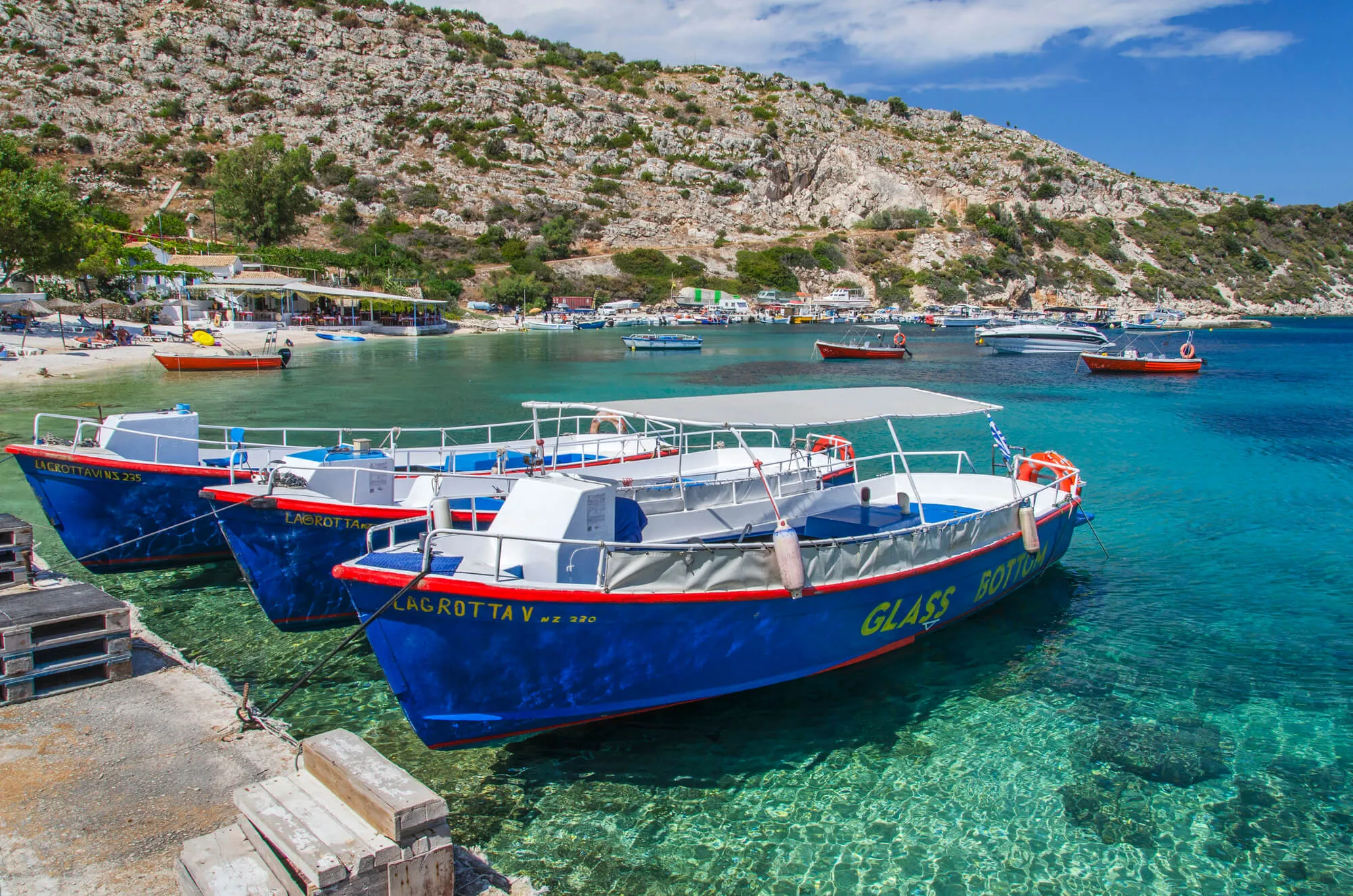 Colorful fishing boats moored in turquoise Mediterranean waters near a rocky hillside village.