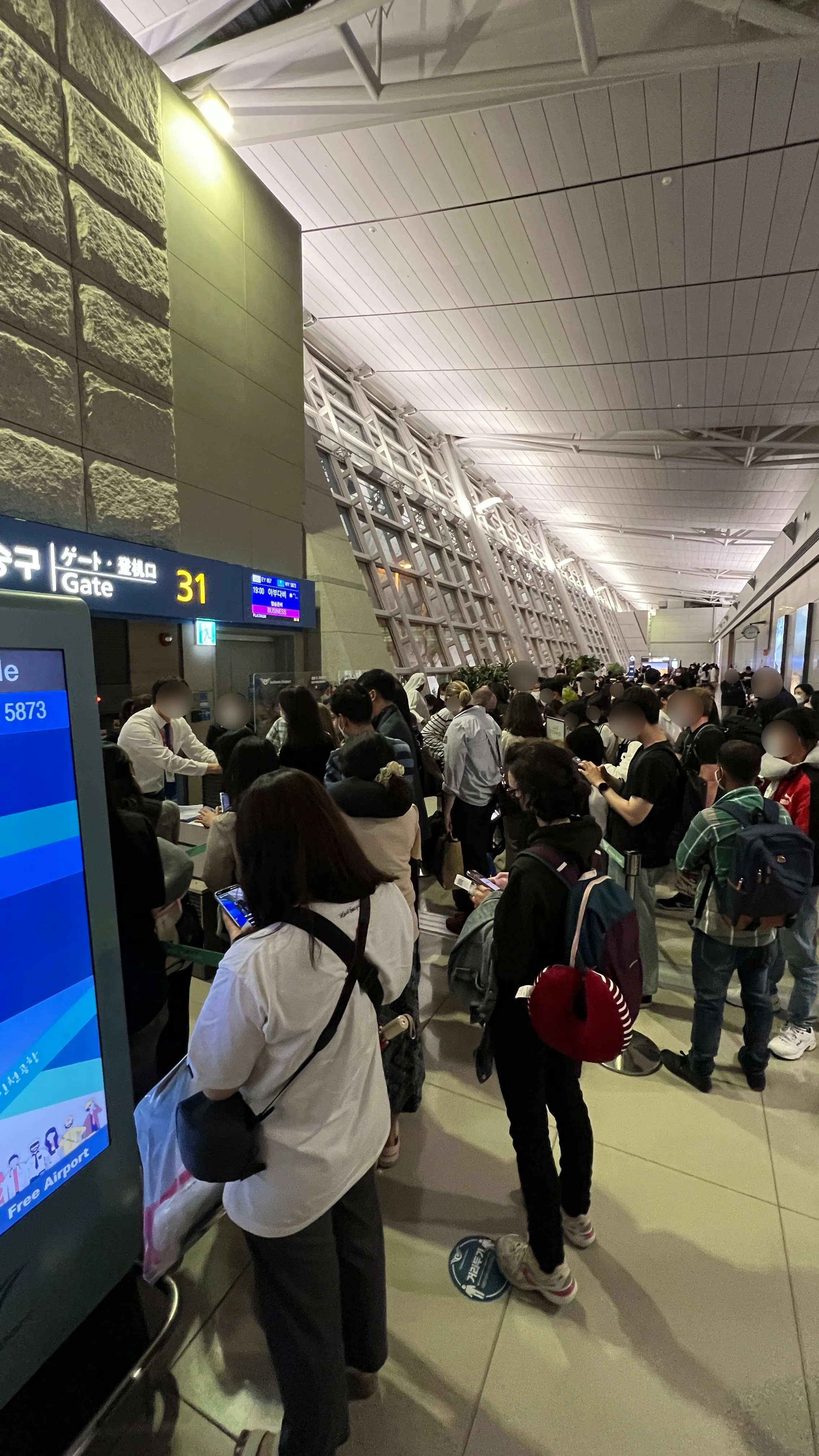 Crowded airport gate area with passengers queuing, Gate 31 sign visible, modern terminal with stone wall accent