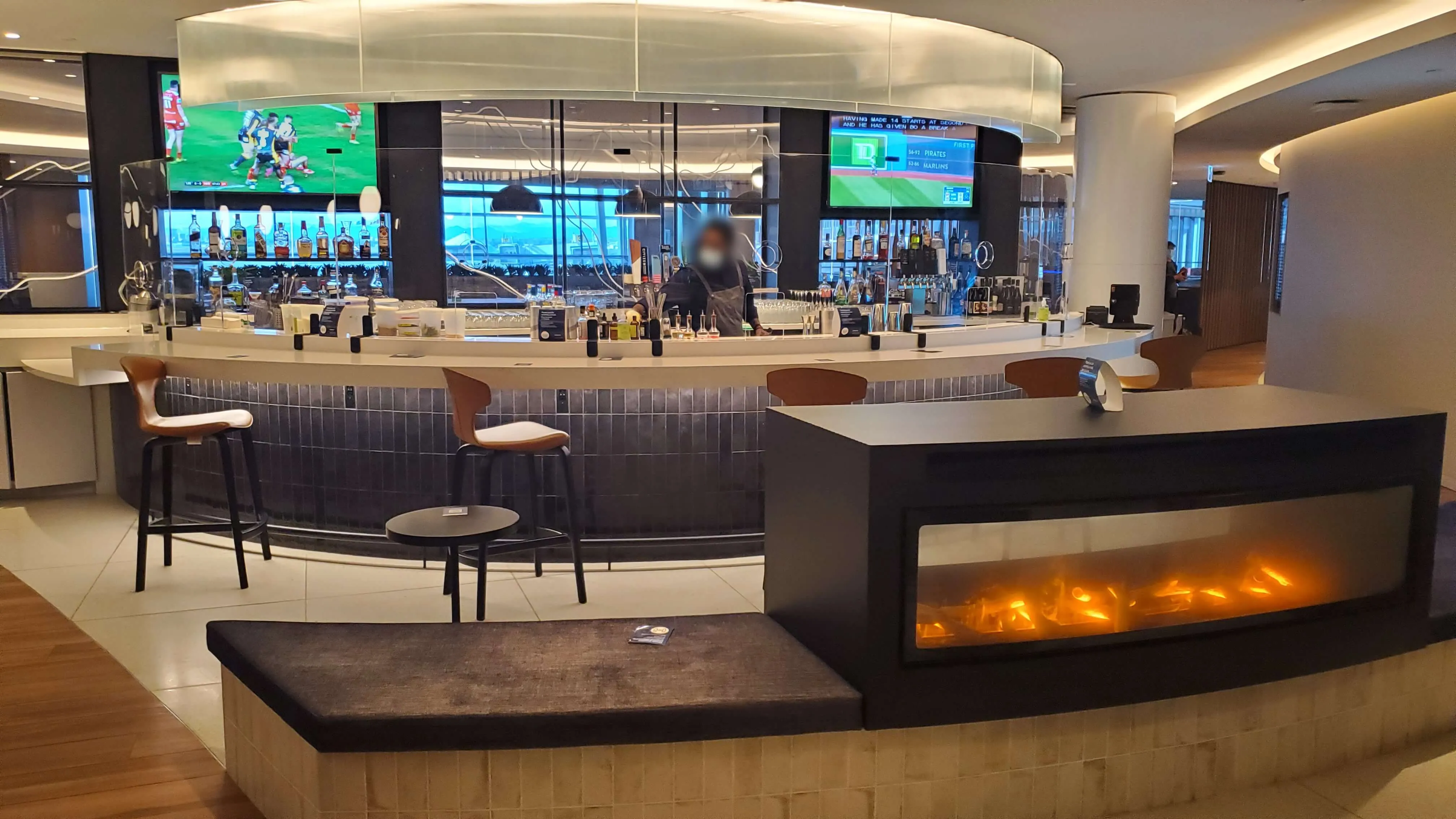 Modern airport lounge with curved bar seating, fireplace feature, and mounted TVs showing sports content, floor-to-ceiling windows overlooking runway