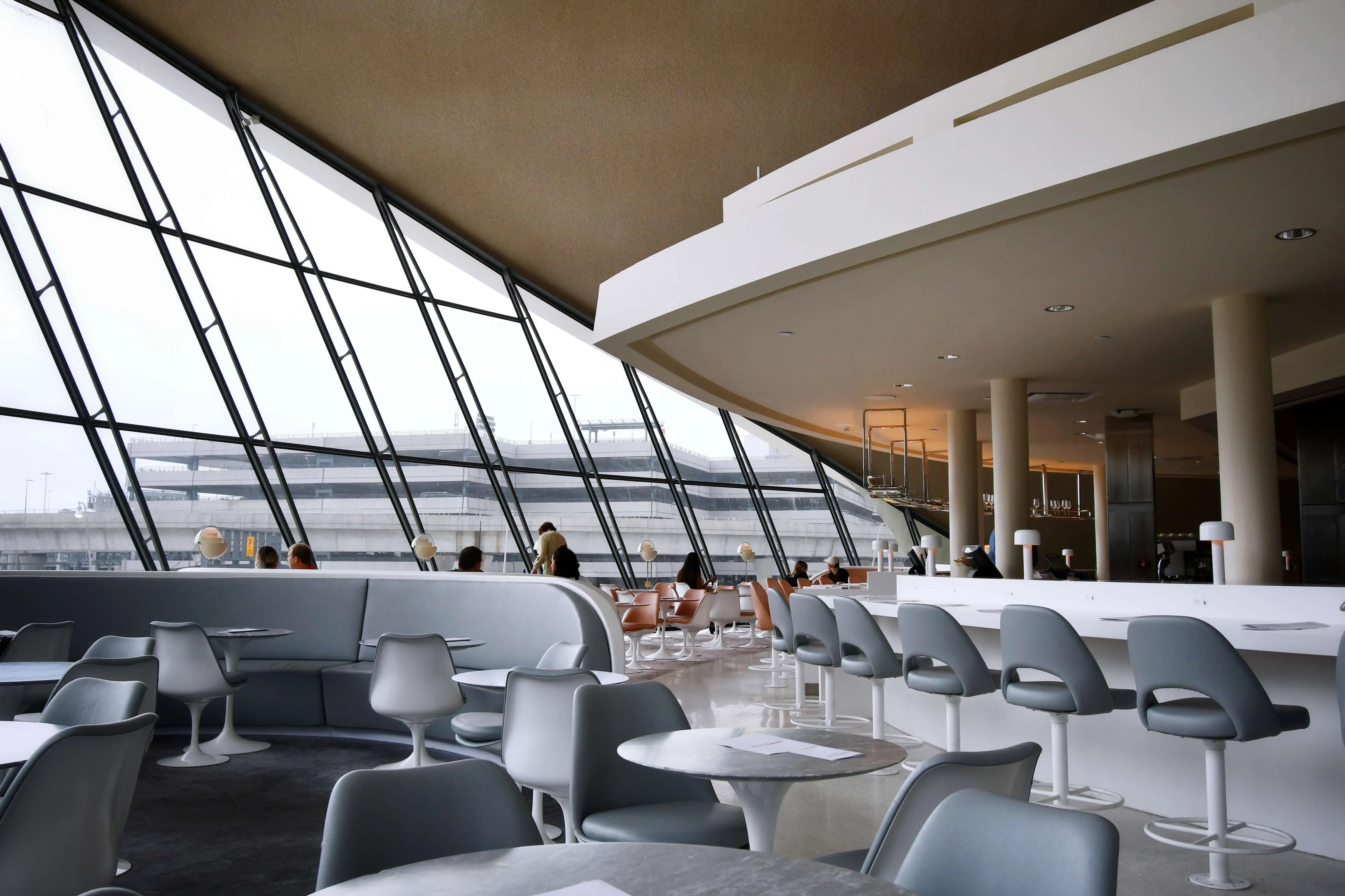 Modern airport lounge with floor-to-ceiling angled windows, gray seating, and tarmac view