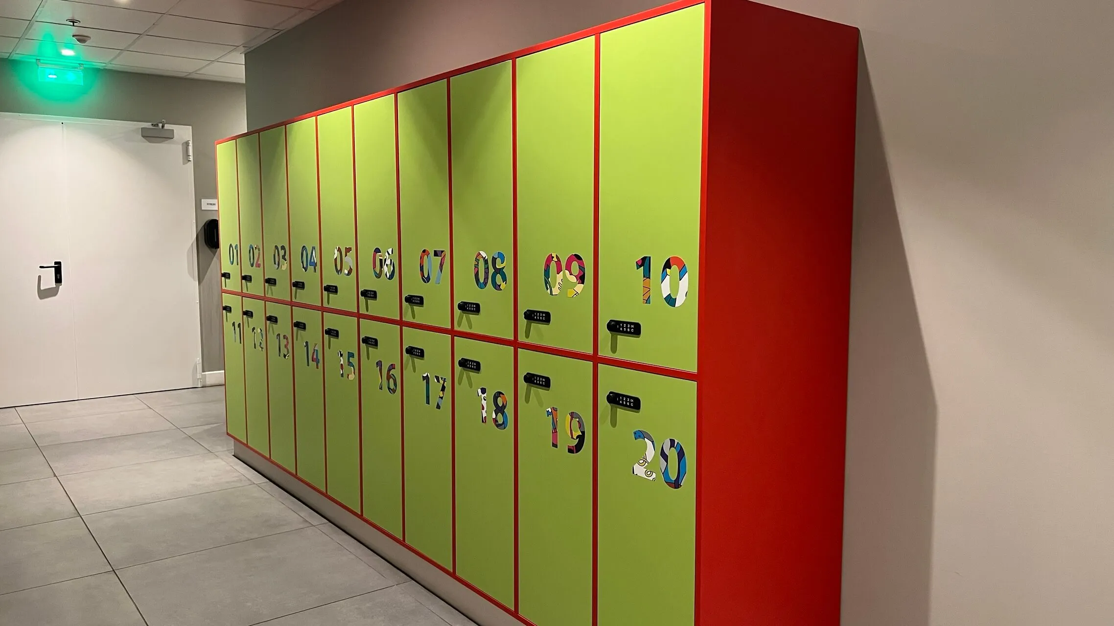 Bright green and red numbered lockers in an airport terminal corridor with tile flooring