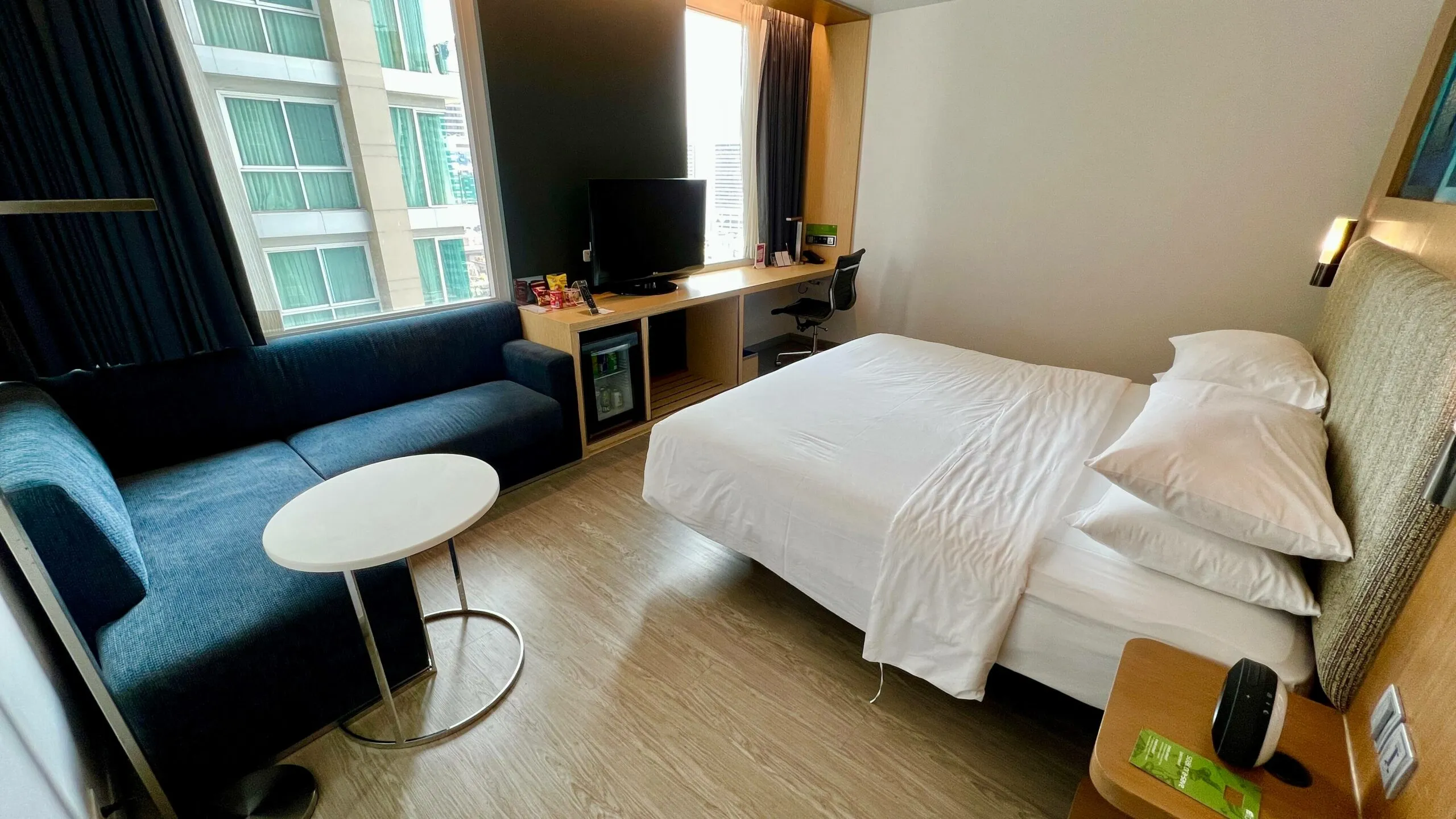 Aloft Bangkok Sukhumvit 11 hotel room with queen bed, blue sofa, TV desk, and floor-to-ceiling windows overlooking the city.