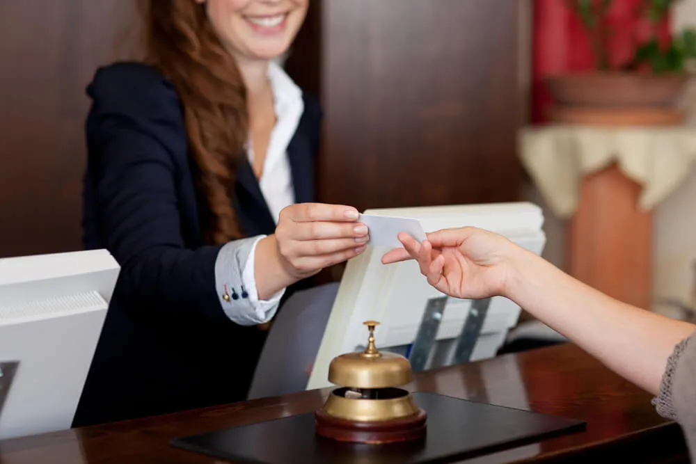 Hotel receptionist handing a key card to a guest at a front desk with a service bell nearby.