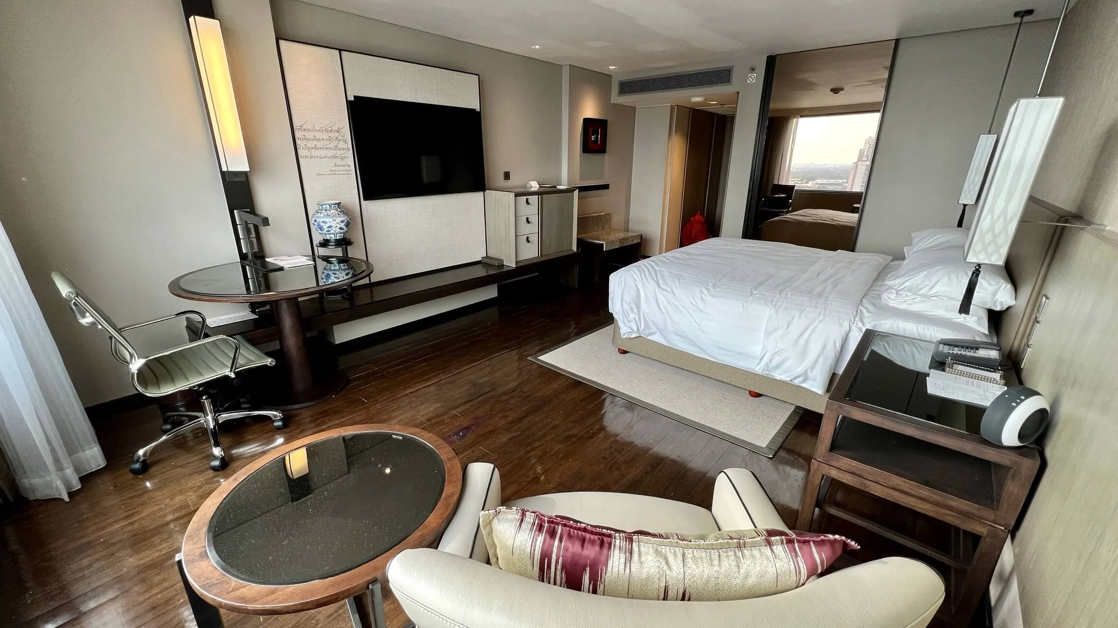 Spacious hotel suite with king bed, living area with curved sofa, work desk, wall-mounted TV, and modern furnishings at Bangkok Marriott Marquis