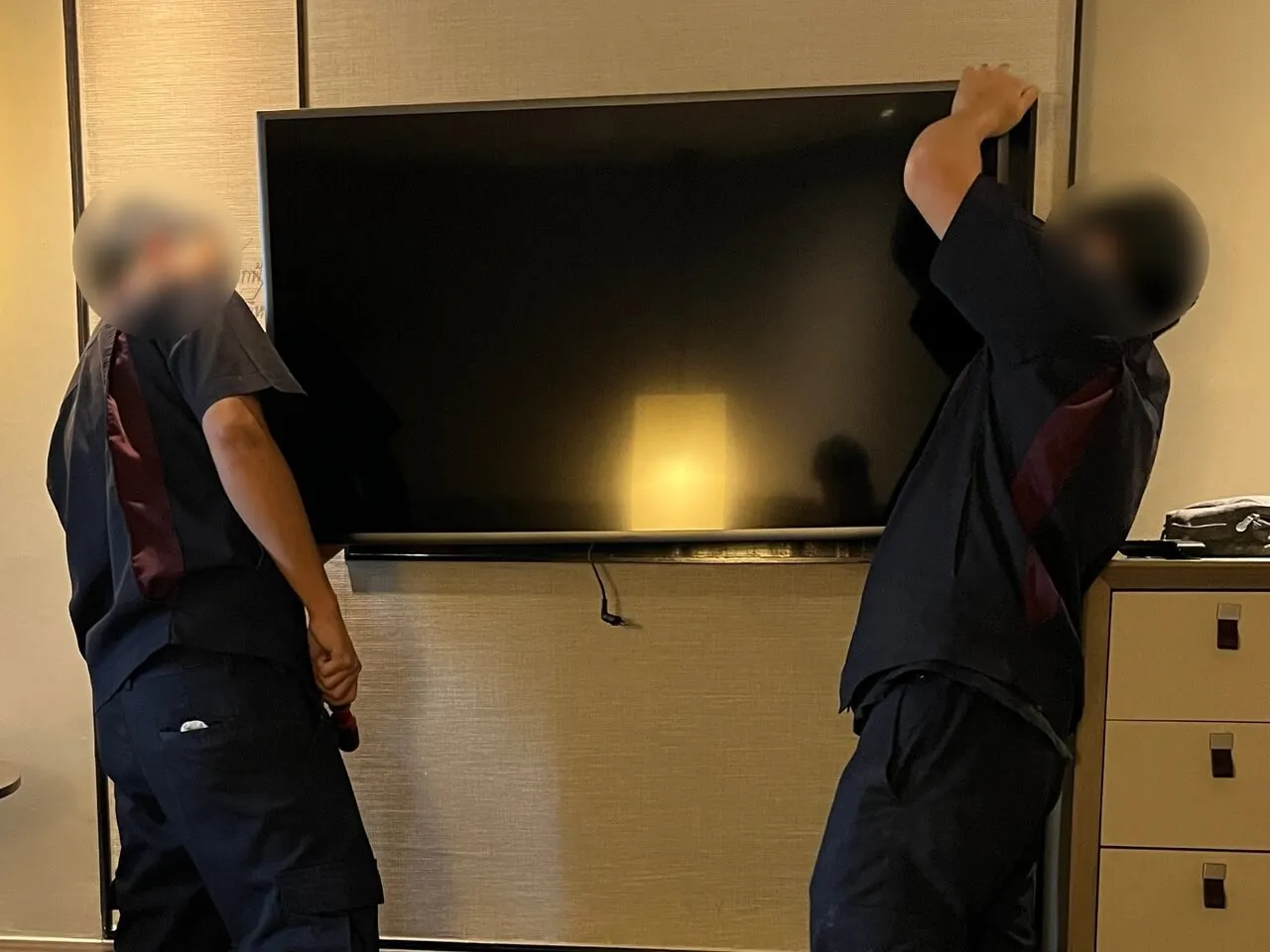 Two maintenance workers adjusting a large flat-screen TV mounted on a wall in a hotel room