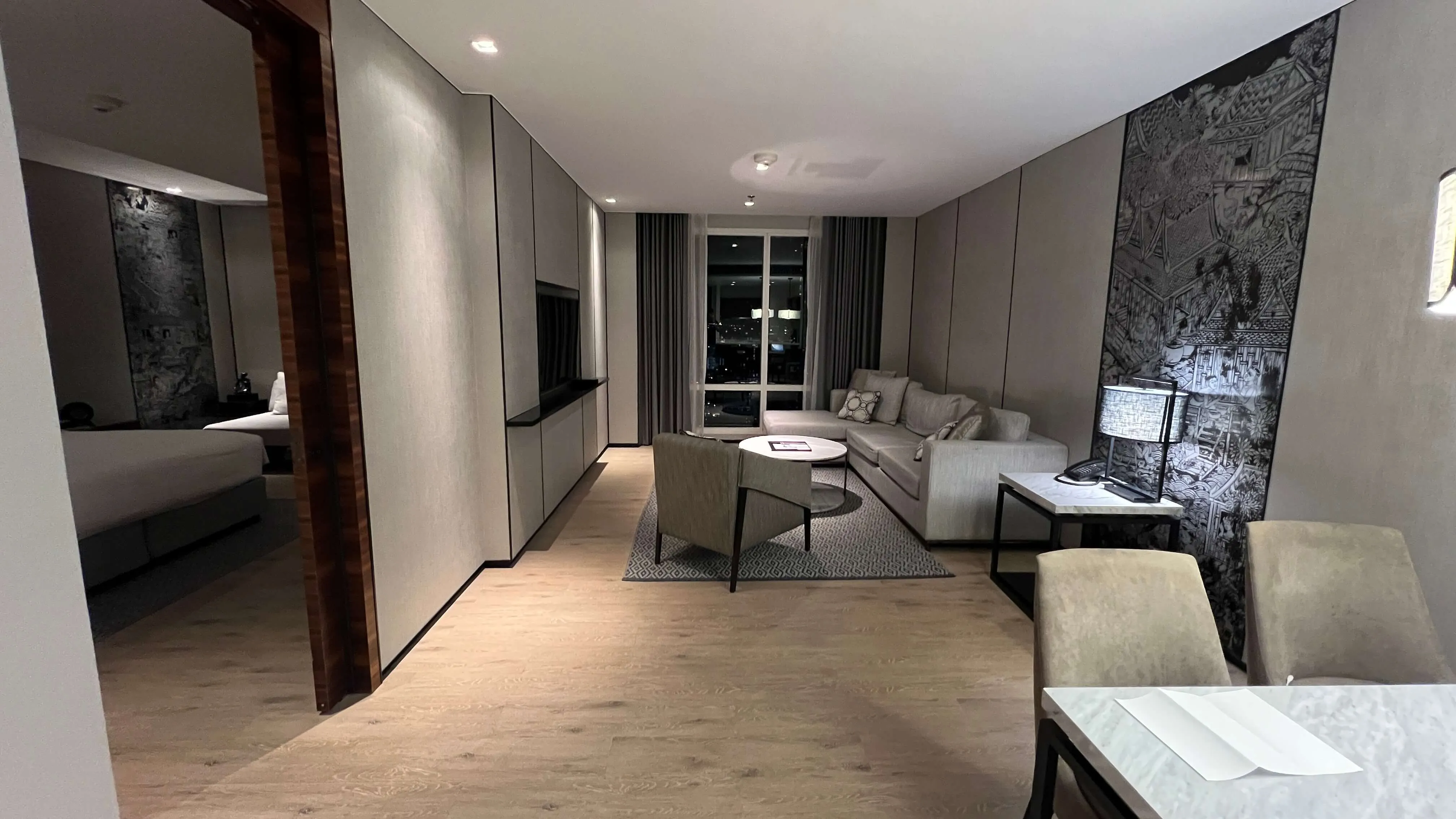 Modern suite with open-plan living area, grey sofa, workspace, wooden flooring, and floor-to-ceiling windows at Marriott Surawongse Bangkok