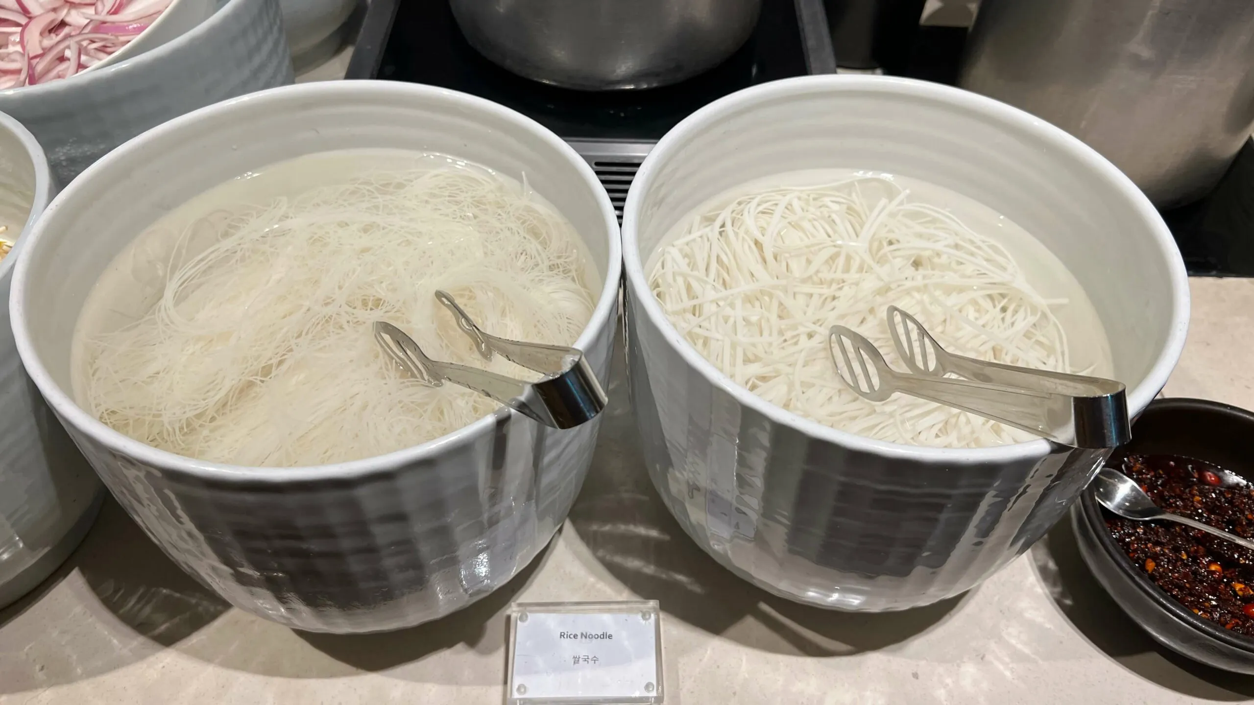 Two white bowls of rice noodles with serving utensils at a buffet station