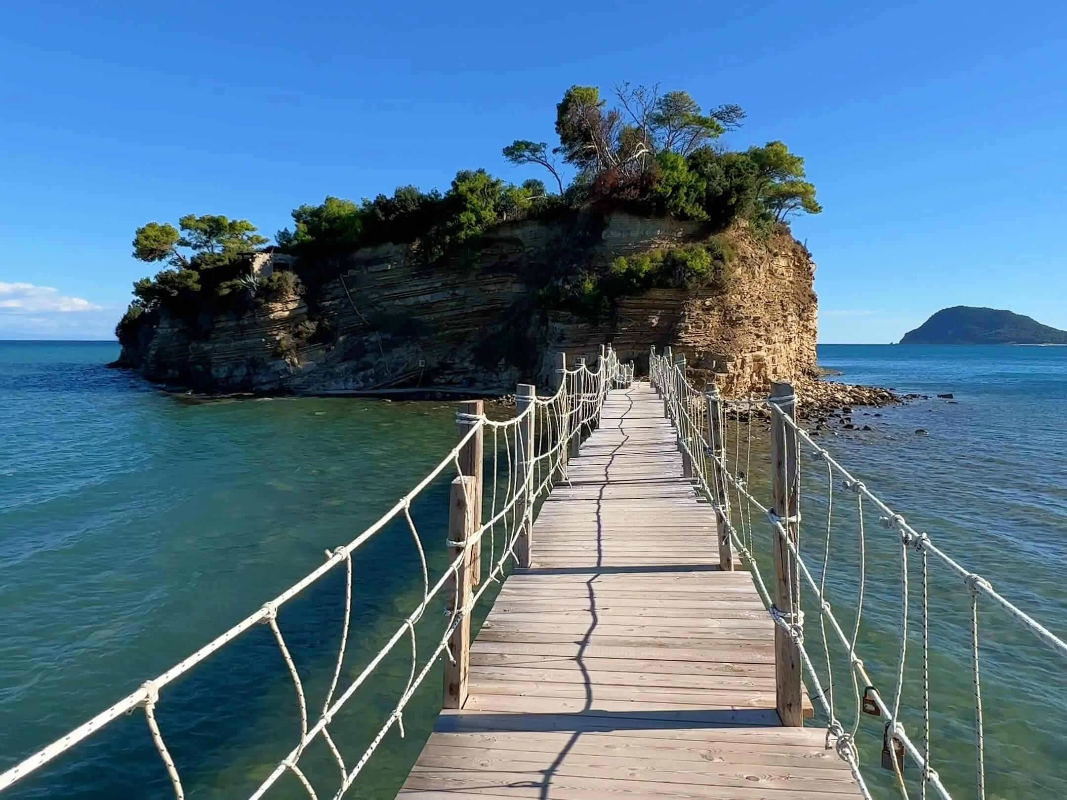 Wooden walkway extending to a rocky island with vegetation under clear blue sky, turquoise water below