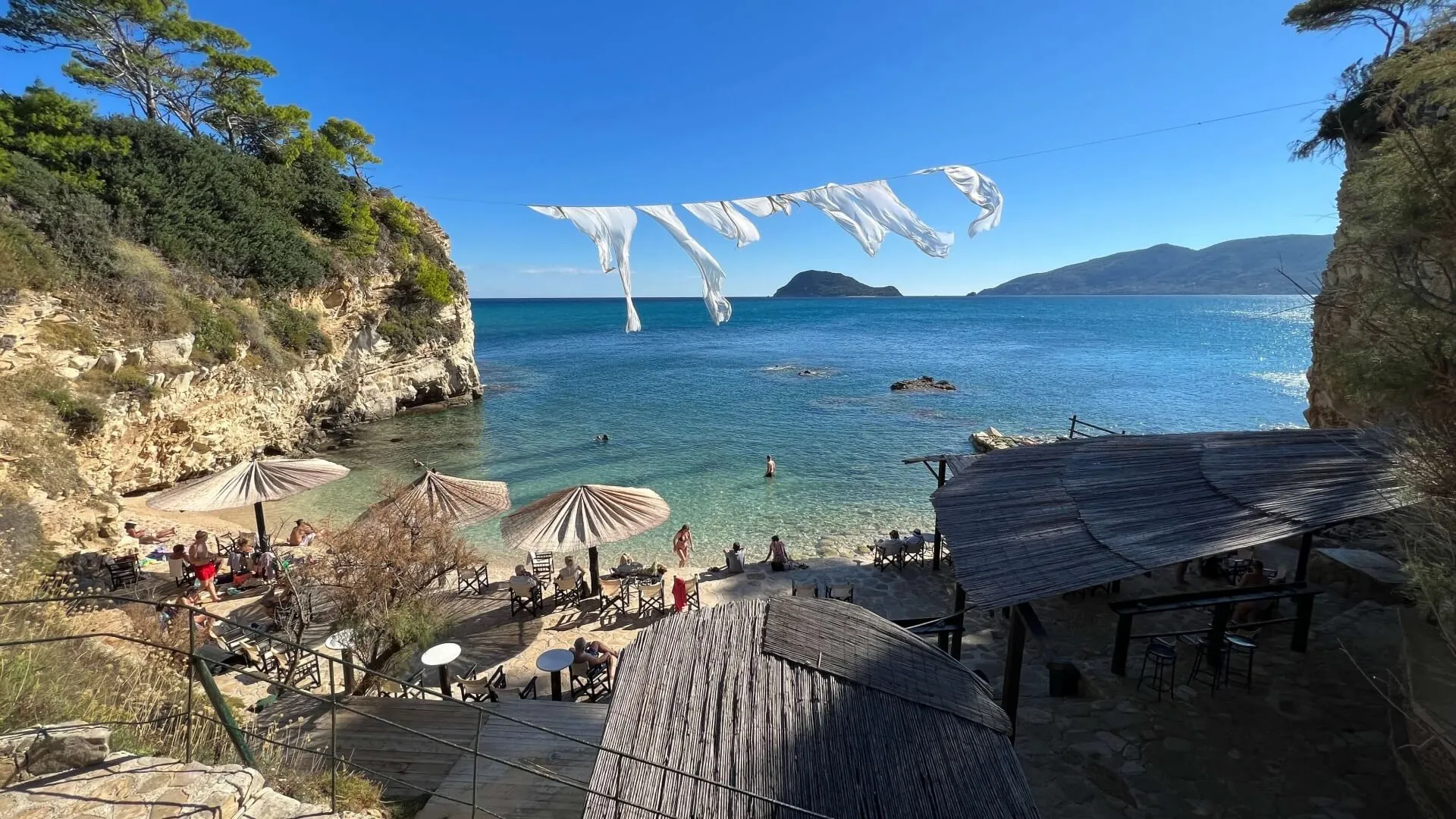 Cove beach with wooden deck, striped umbrellas, turquoise Aegean water, and sailboat in distance framed by limestone cliffs