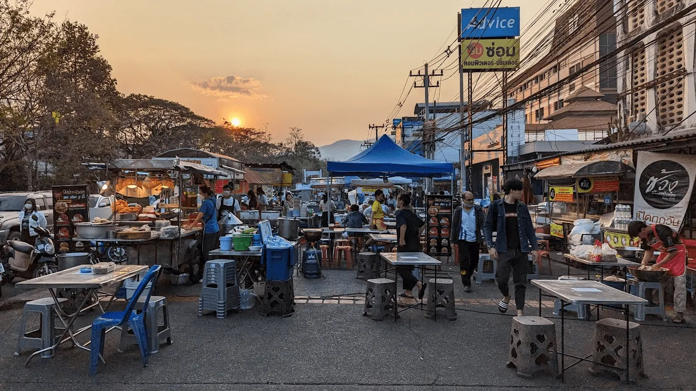 Street food vendors and diners at Chang Phuak night market at sunset, with blue signage and mountain backdrop