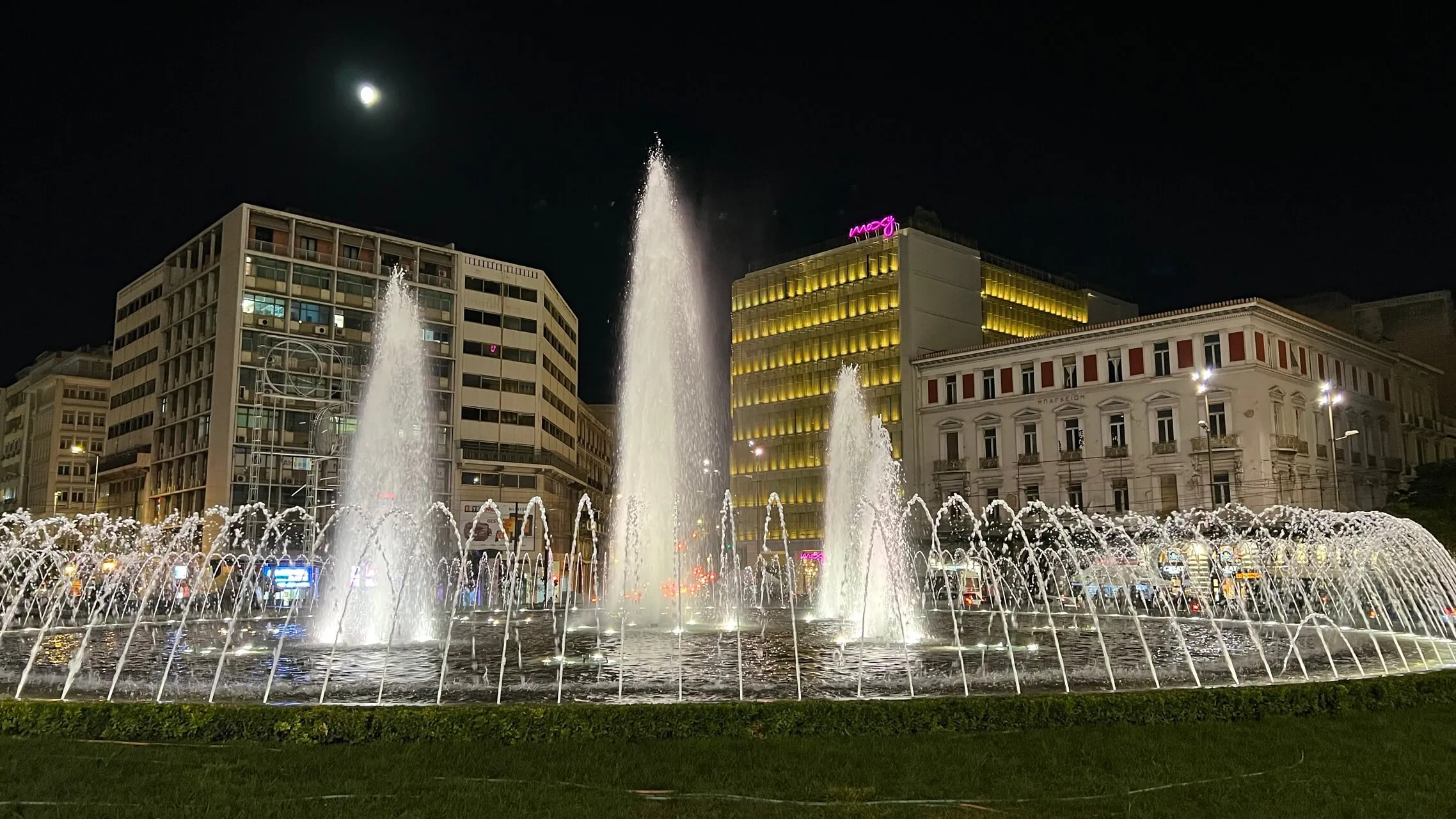 Illuminated fountains in a city square at night surrounded by modern and classical buildings under moonlight