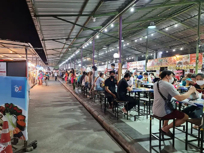 Busy outdoor night market with food stalls, vendor booths, and diners seated at tables under metal roof structure