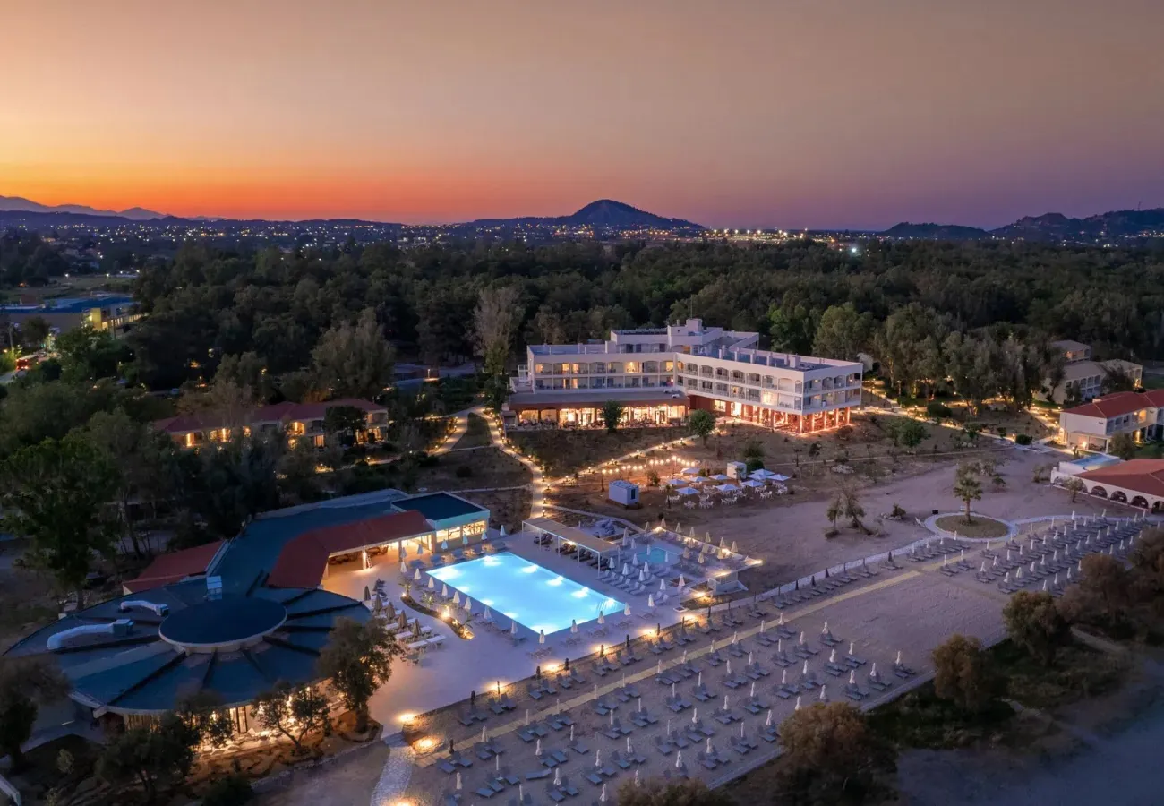 Domes Aulus Zante Autograph Collection resort at dusk with illuminated pool, beach loungers, and main building overlooking forested landscape and distant mountains