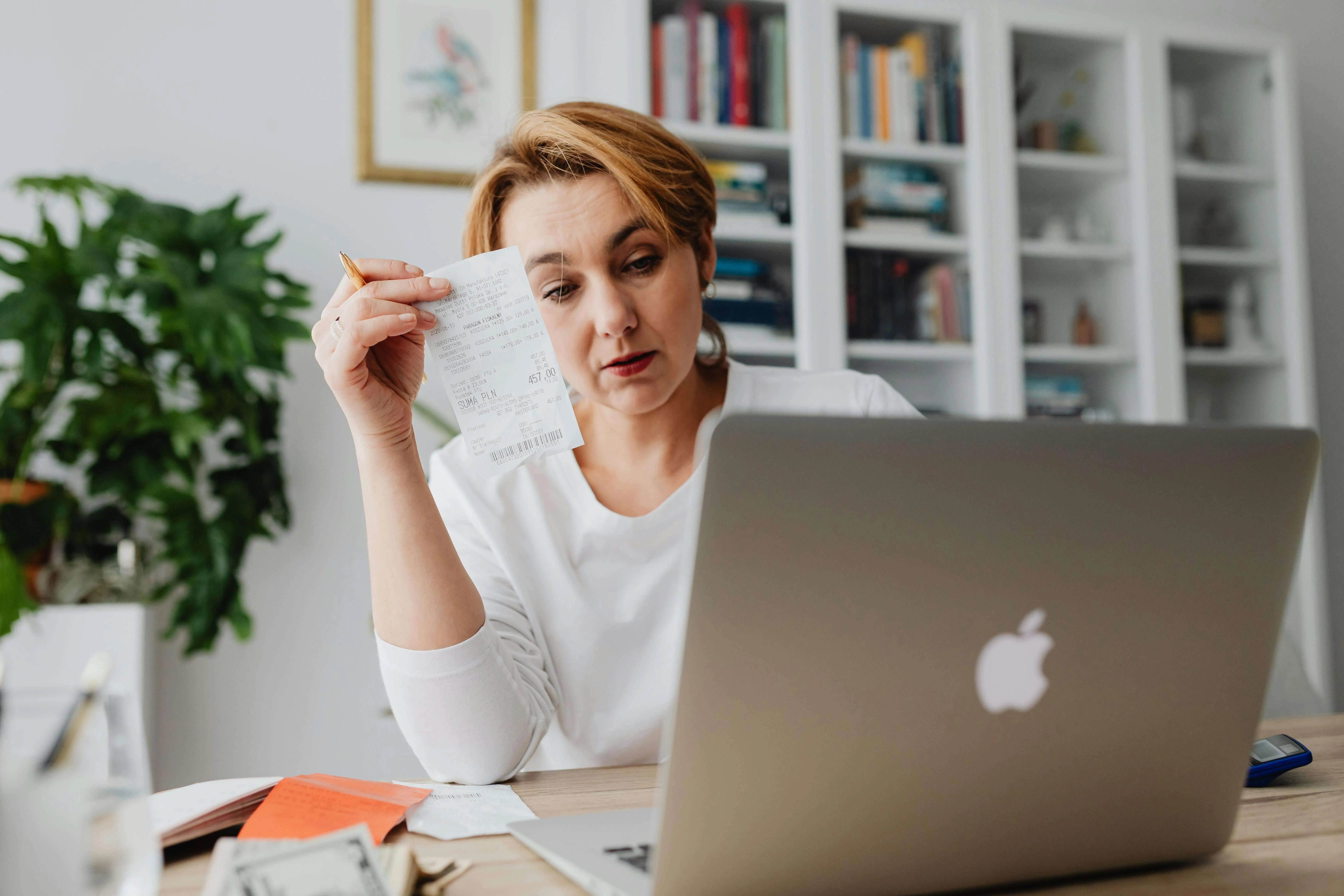 Woman at desk with MacBook laptop reviewing receipt, surrounded by white shelving and plants