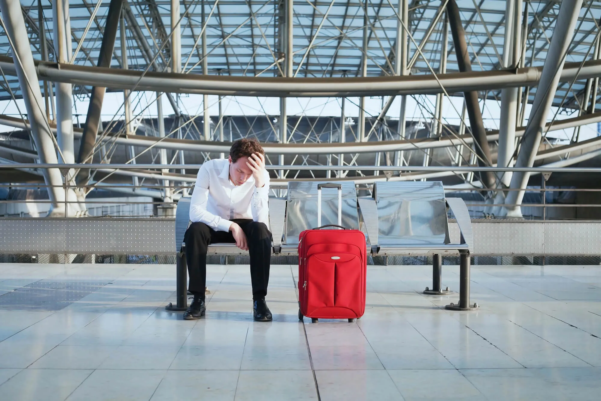 Traveler sitting on airport bench with red luggage, appearing stressed or tired