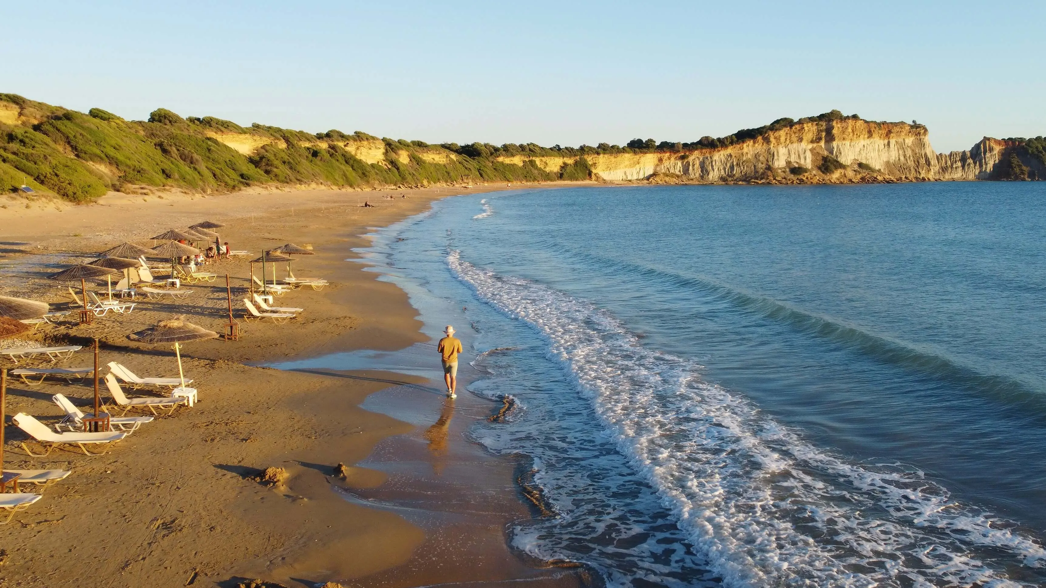 Gerakas Beach with golden sand, beach umbrellas and loungers, swimmers in blue Aegean waters, and limestone cliffs in the background