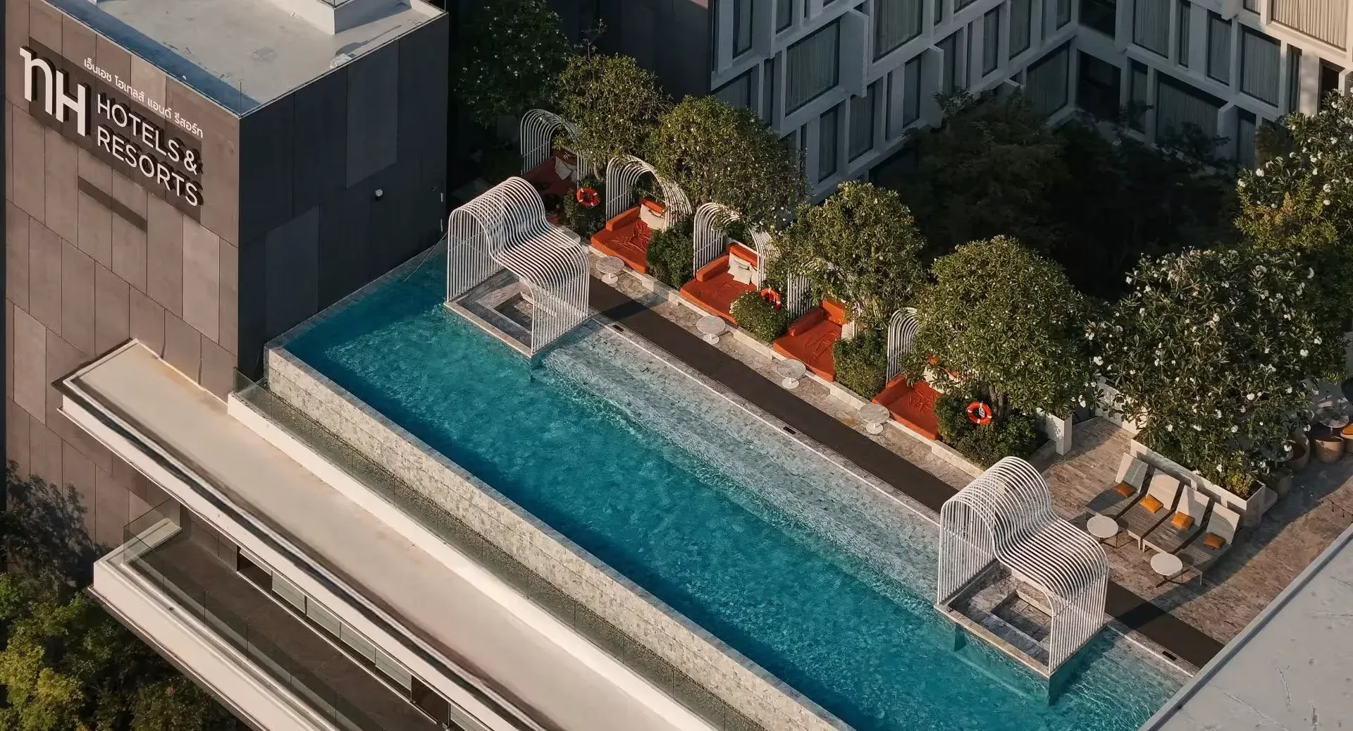 NH Hua Hin's pool deck from above: a long turquoise pool with white arched cabanas, orange daybeds under frangipani trees and the NH Hotels & Resorts sign on the tower