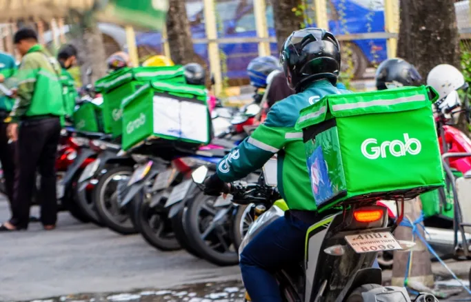 Grab delivery driver on motorcycle with green insulated food bag in urban street setting with other riders.