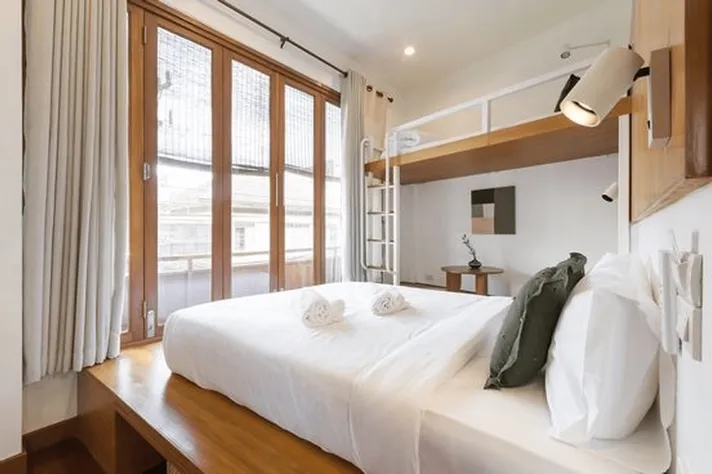 Contemporary hostel room with queen bed, wooden flooring, large windows with curtains, and lofted sleeping area above.