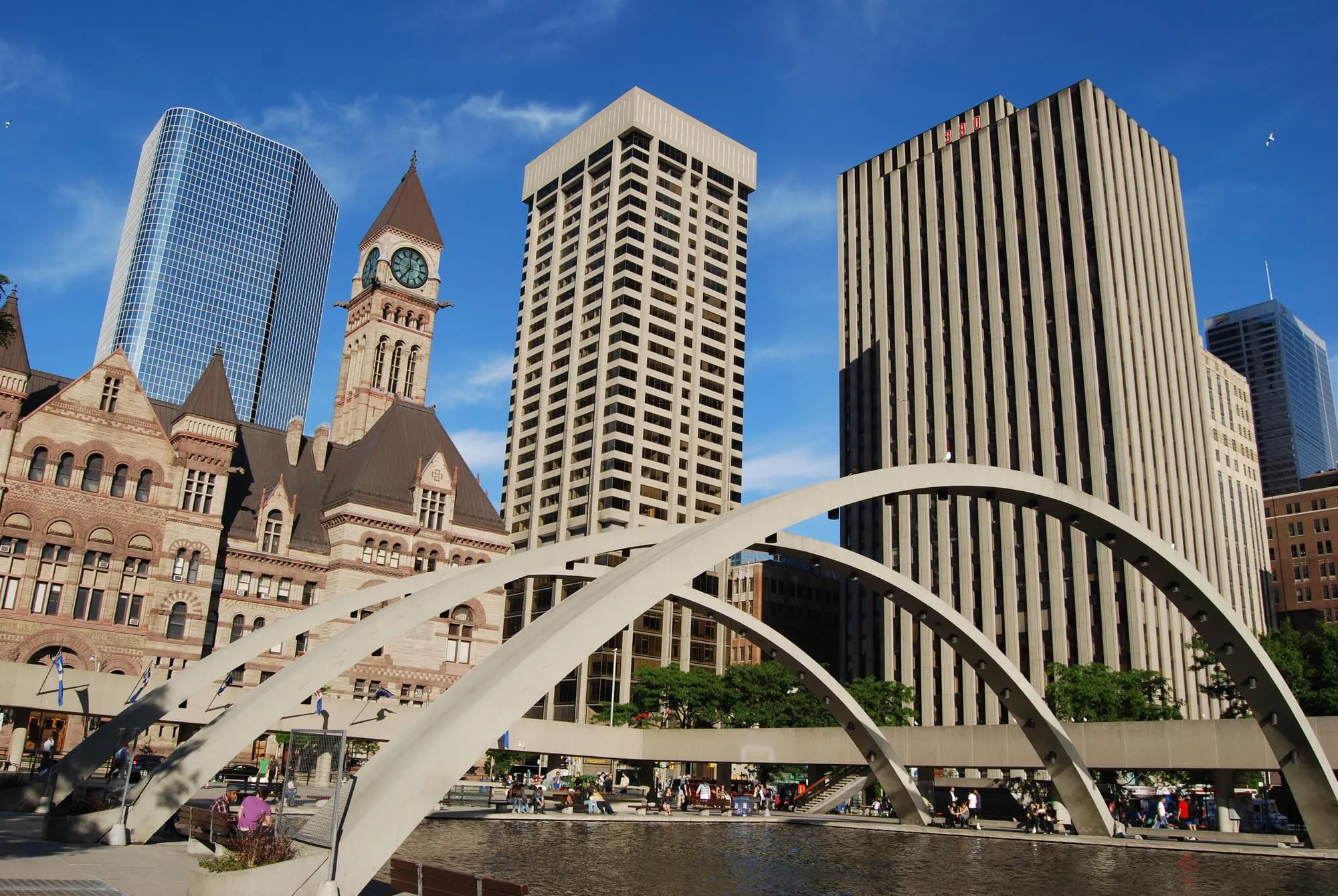 Toronto City Hall with clock tower, modern high-rises, and white arched bridge over Nathan Phillips Square, clear blue sky