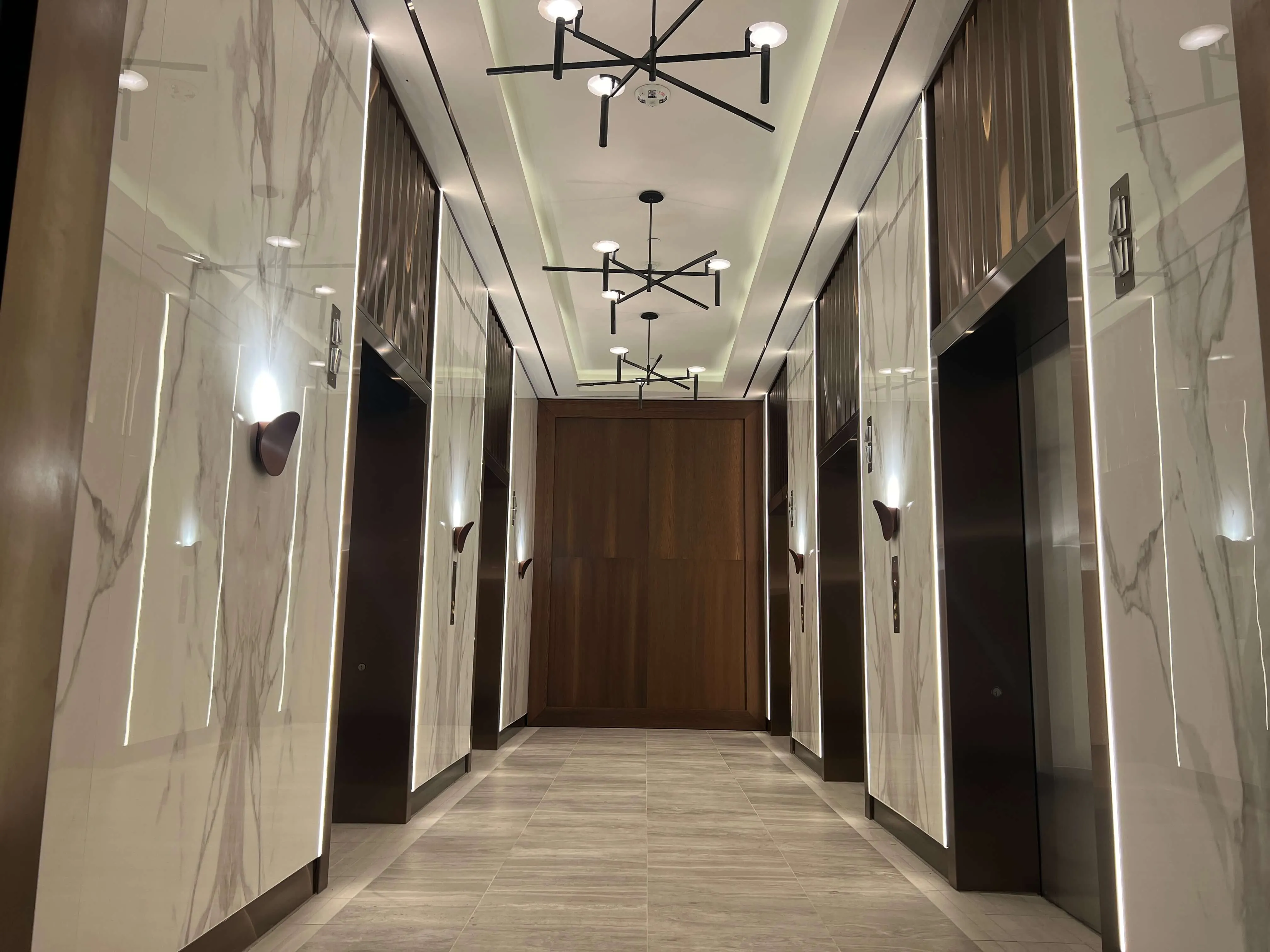 Modern hotel corridor with marble-paneled walls, contemporary black pendant lights, and elevator doors on either side.