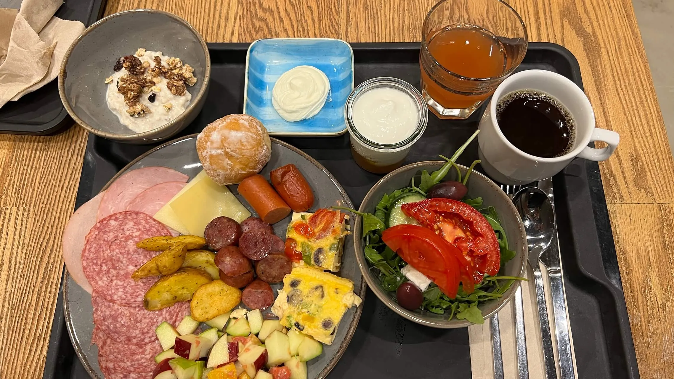 Continental breakfast spread with cured meats, cheese, pastry, roasted potatoes, frittata, fresh salad, yogurt, juices, and coffee on a black tray.