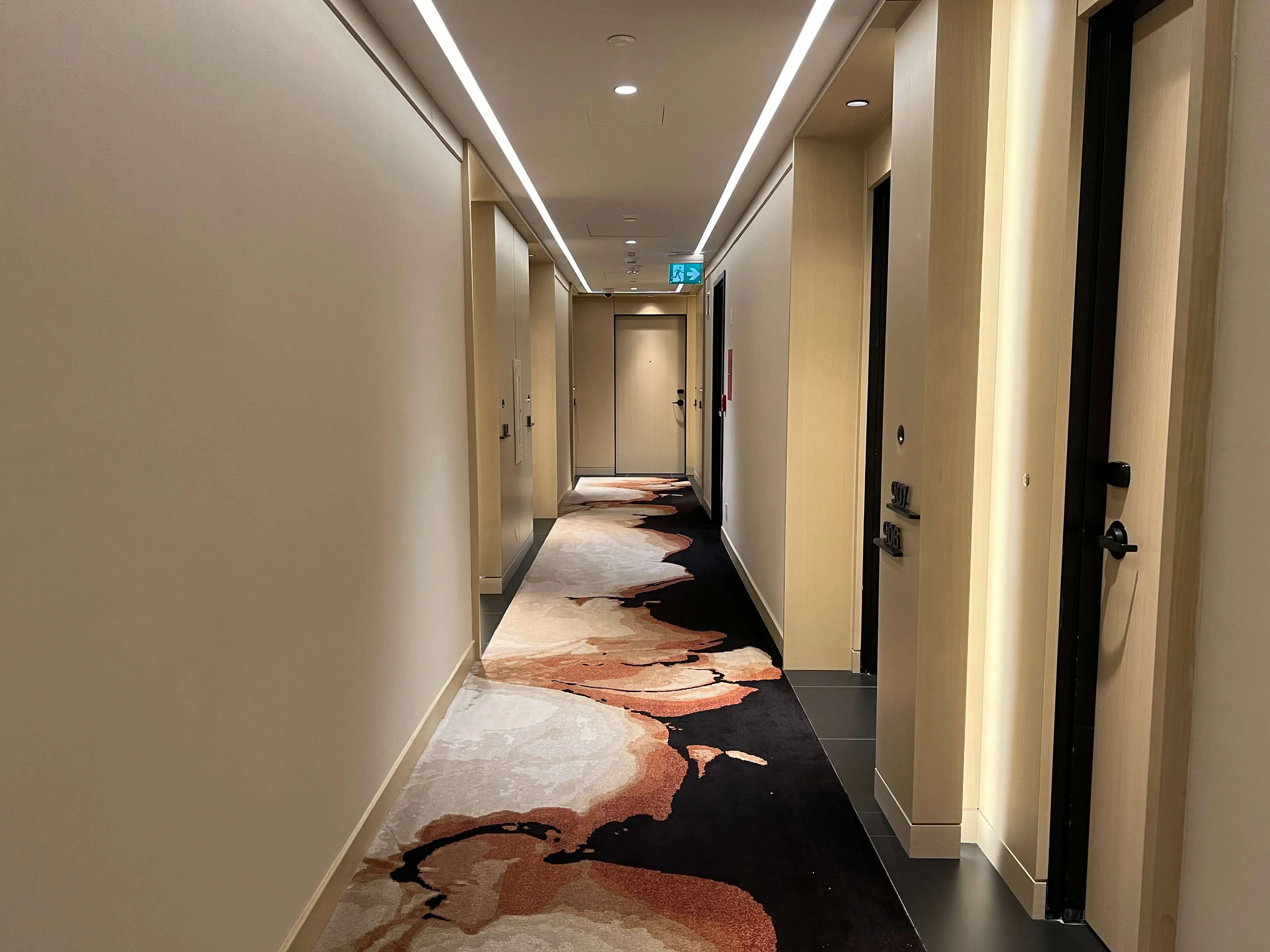 Hotel corridor with cream walls, linear ceiling lights, and a black floor featuring abstract rust and cream watercolor-style artwork.