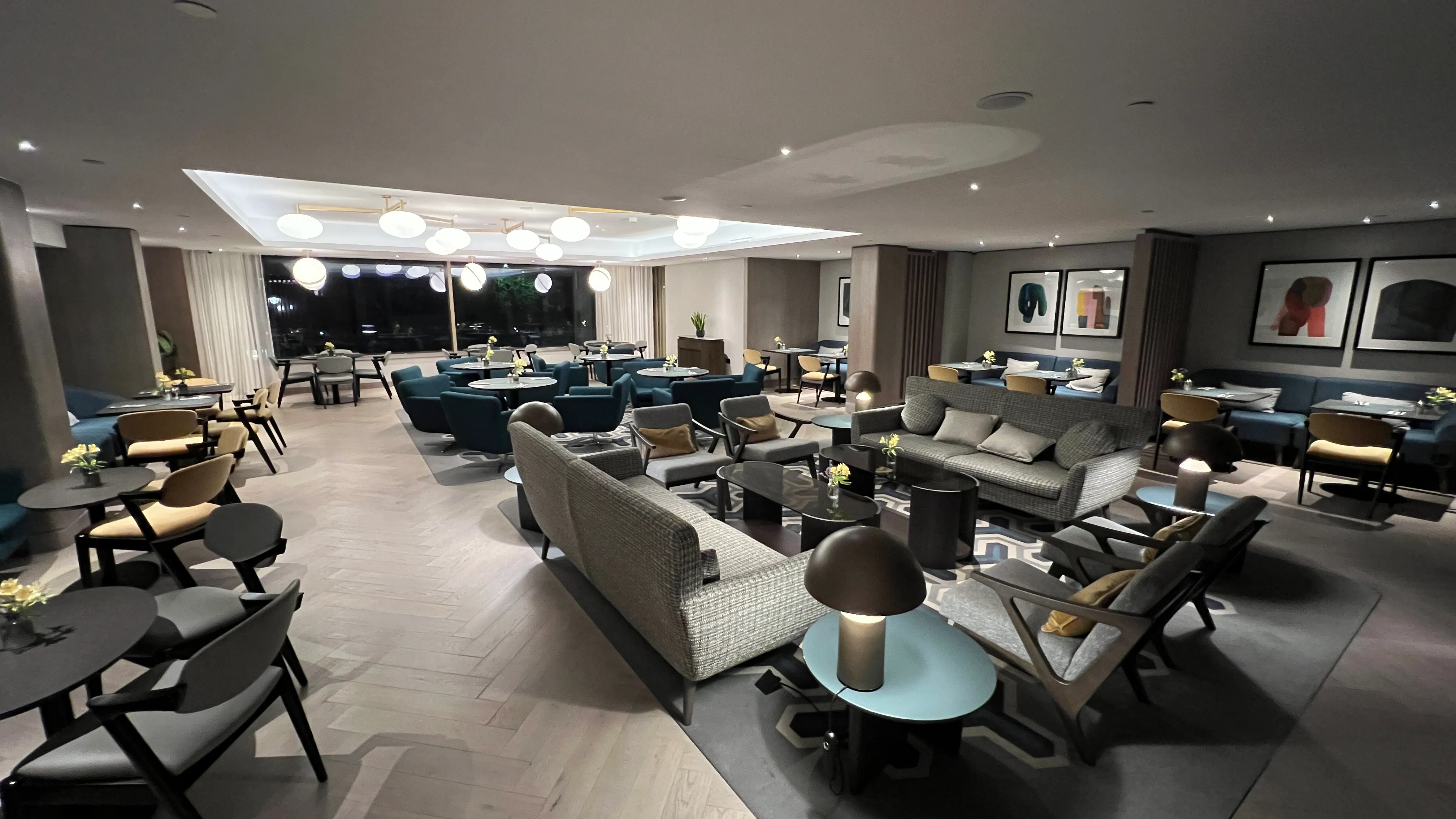 Modern hotel lounge with blue and gray seating, pendant lighting, and floor-to-ceiling windows overlooking a city at night