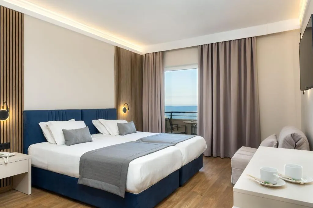Modern hotel bedroom with navy upholstered bed, sea view from balcony, cove ceiling lighting, and contemporary furnishings