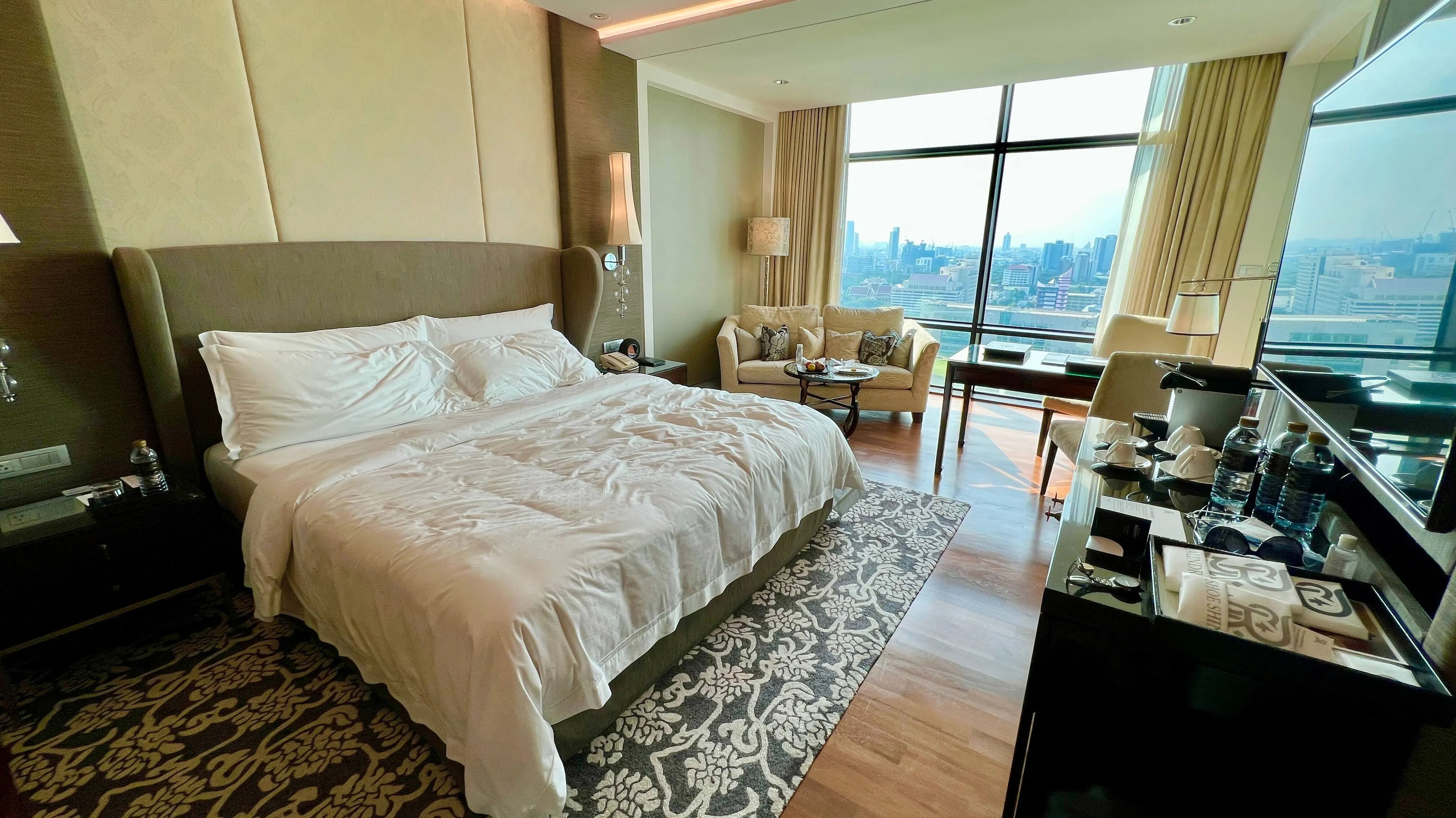 Spacious hotel room with king bed, city skyline view through floor-to-ceiling windows, sitting area, and modern furnishings.