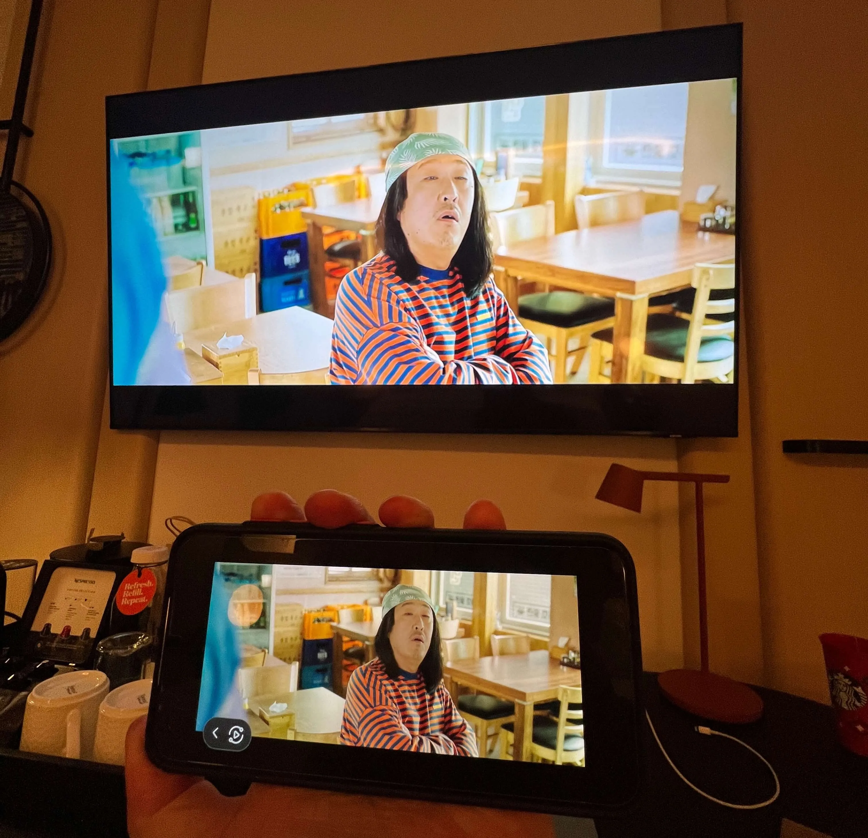 TV screen and tablet displaying a woman in a striped shirt and headband in a bright, modern dining space with wooden tables and city views