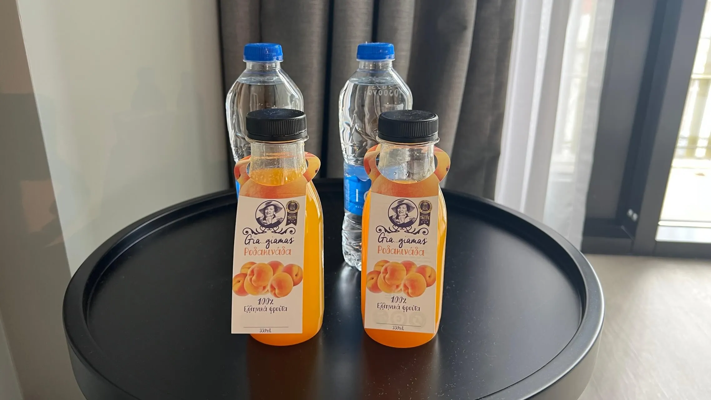 Complimentary bottled juices on a dark round table in a hotel room, with curtains and window in background