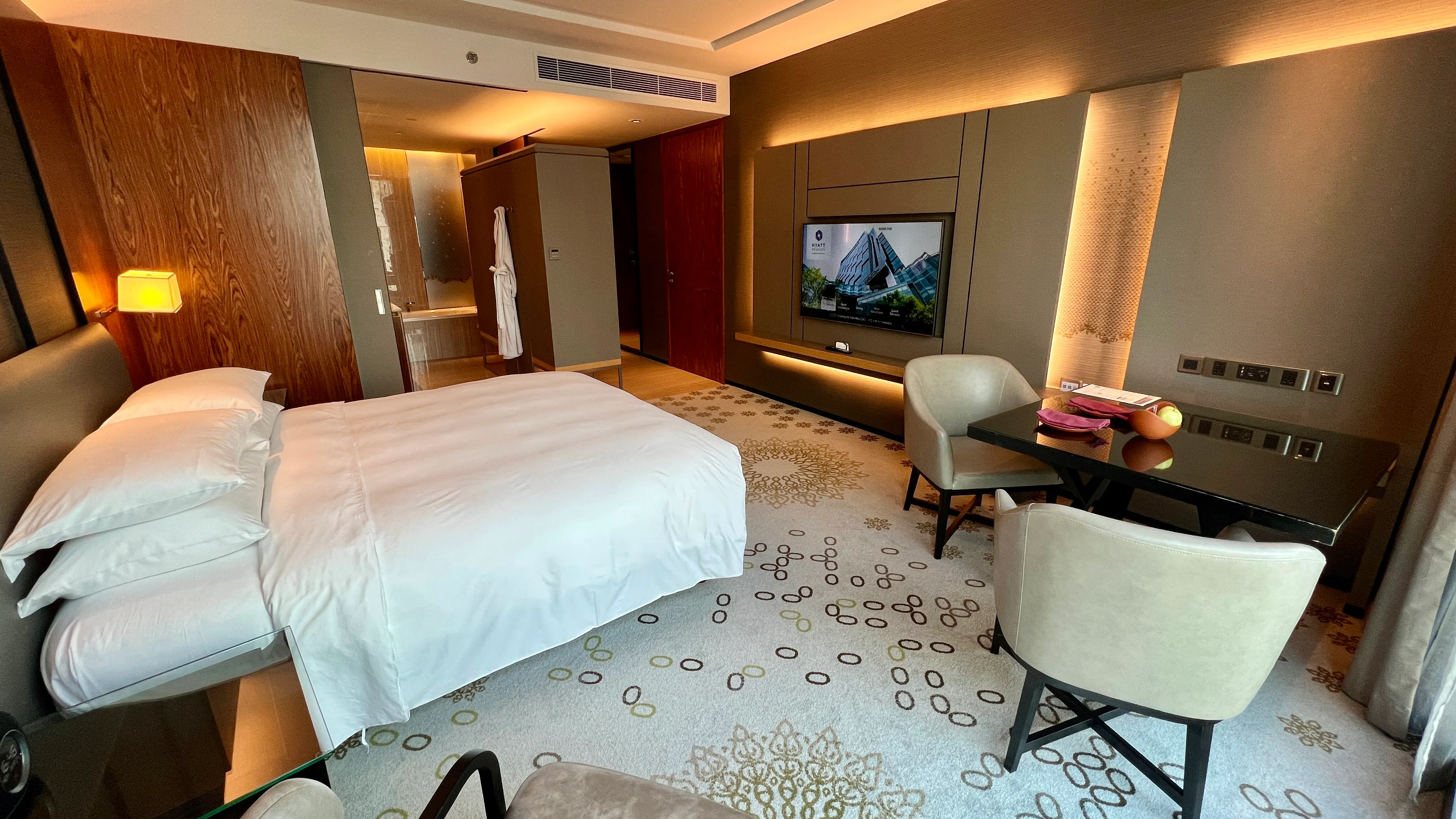 King bed with white linens in warm-lit hotel room at Hyatt Regency Bangkok, featuring dark wood paneling, wall-mounted TV, and modern furnishings.