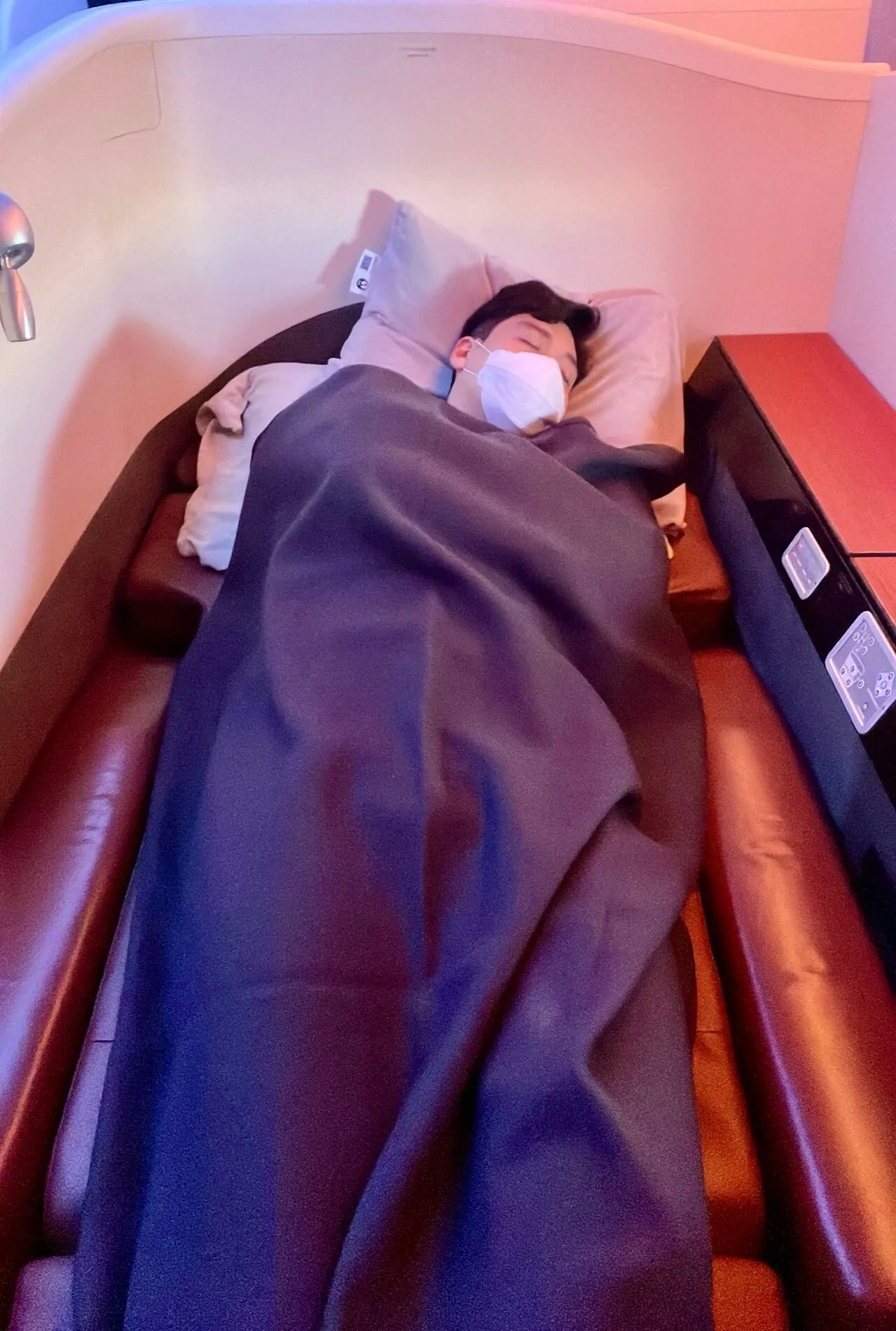 Passenger lying flat in JAL first-class cabin bed with navy blanket and pillow