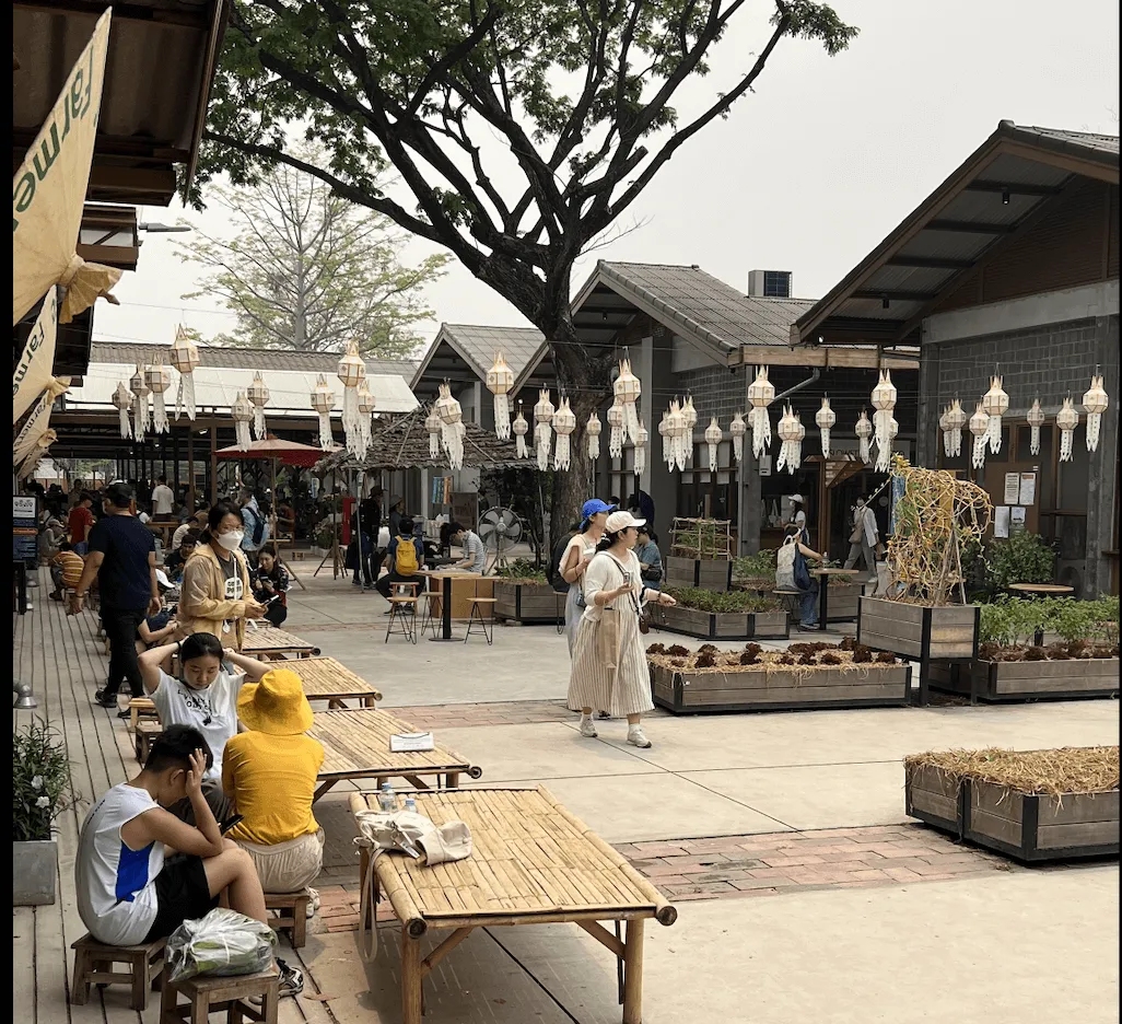 Outdoor courtyard market with wooden picnic tables, hanging white lanterns, traditional gray buildings, and visitors shopping at stalls