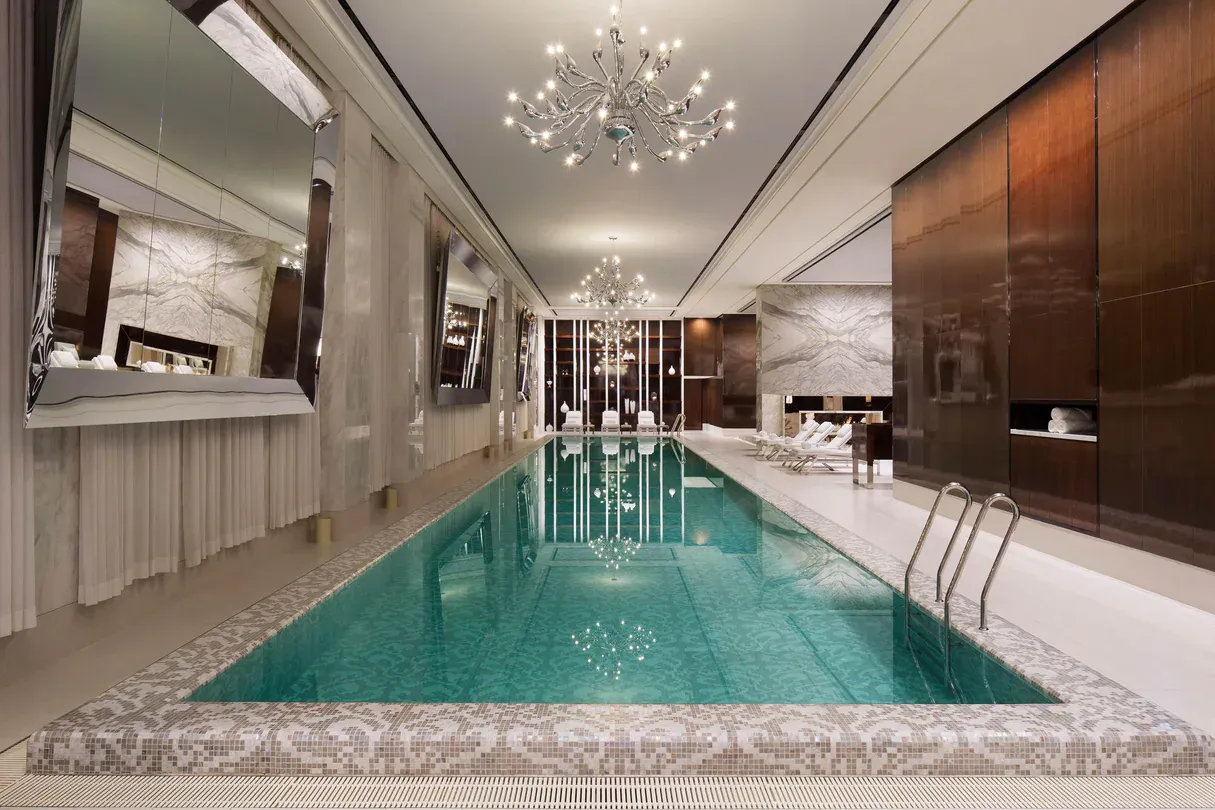 Indoor swimming pool with turquoise water at a luxury hotel, featuring marble and wood finishes, chandeliers, and spa facilities.