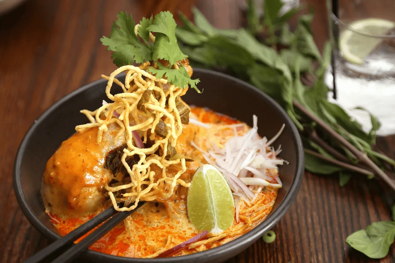 Khao soi noodle curry soup in black bowl with crispy noodles, cilantro, lime, and chopsticks on wooden table